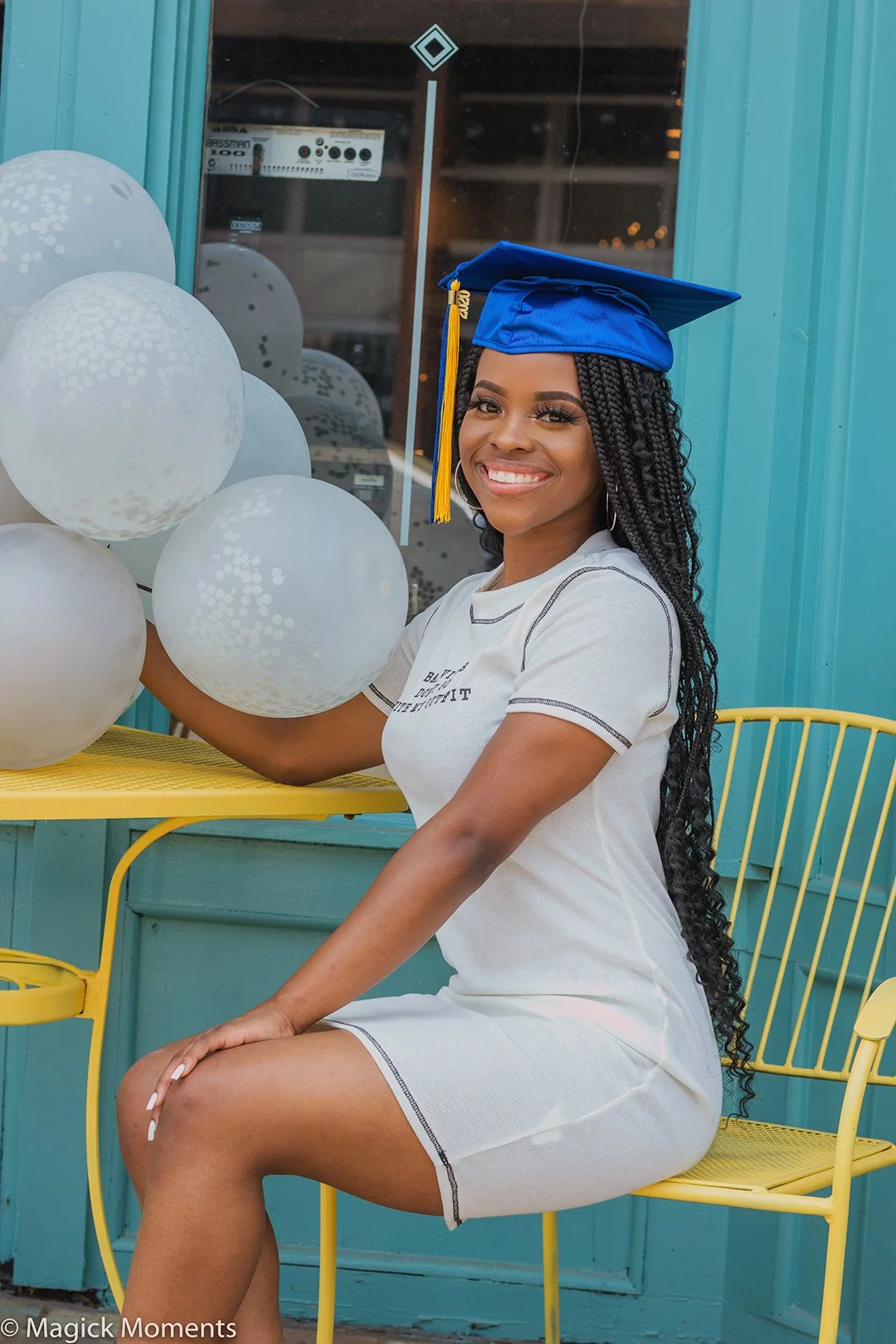 A young woman in a white dress and blue graduation cap sitting on a yellow metal chair. She is smiling and holding white balloons. The background is a teal-colored wall with a window behind her.