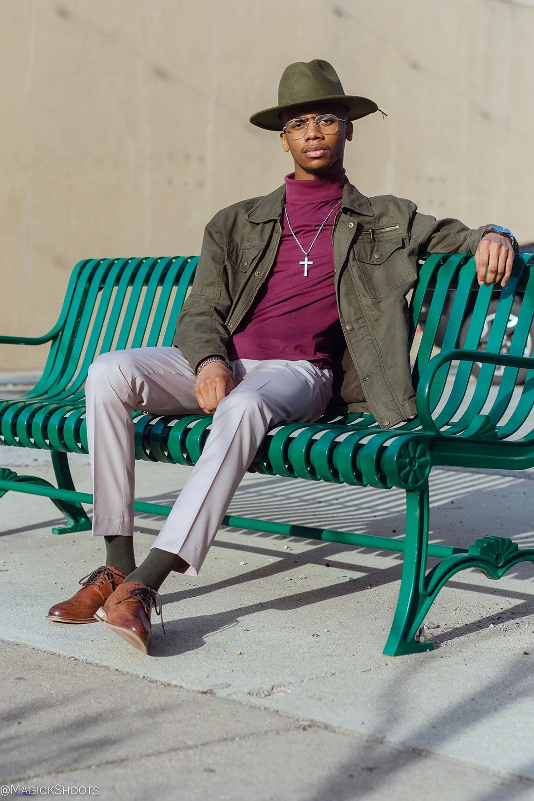 Man sitting on a green park bench outdoors, wearing a wide-brimmed hat, glasses, a dark jacket, a maroon turtleneck, light-colored trousers, and brown shoes. He has a necklace with a cross pendant and a watch.