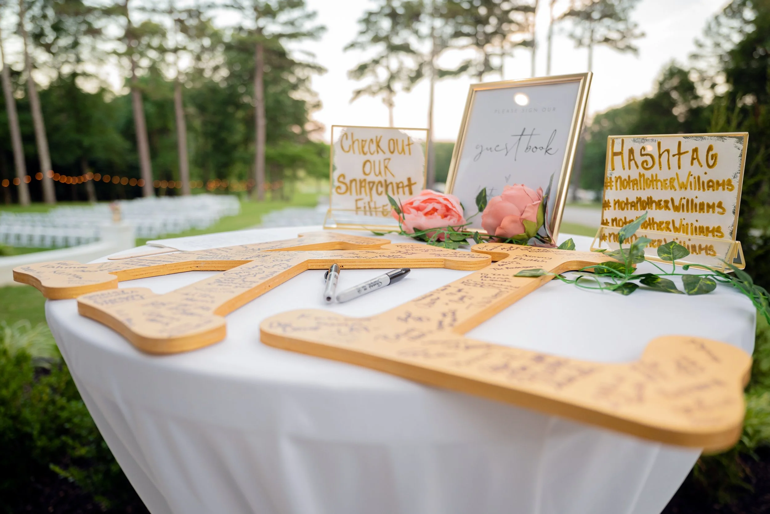 Decorative outdoor table with guest book, framed signs, and floral accents at a wedding or event in a lush, green setting during sunset.