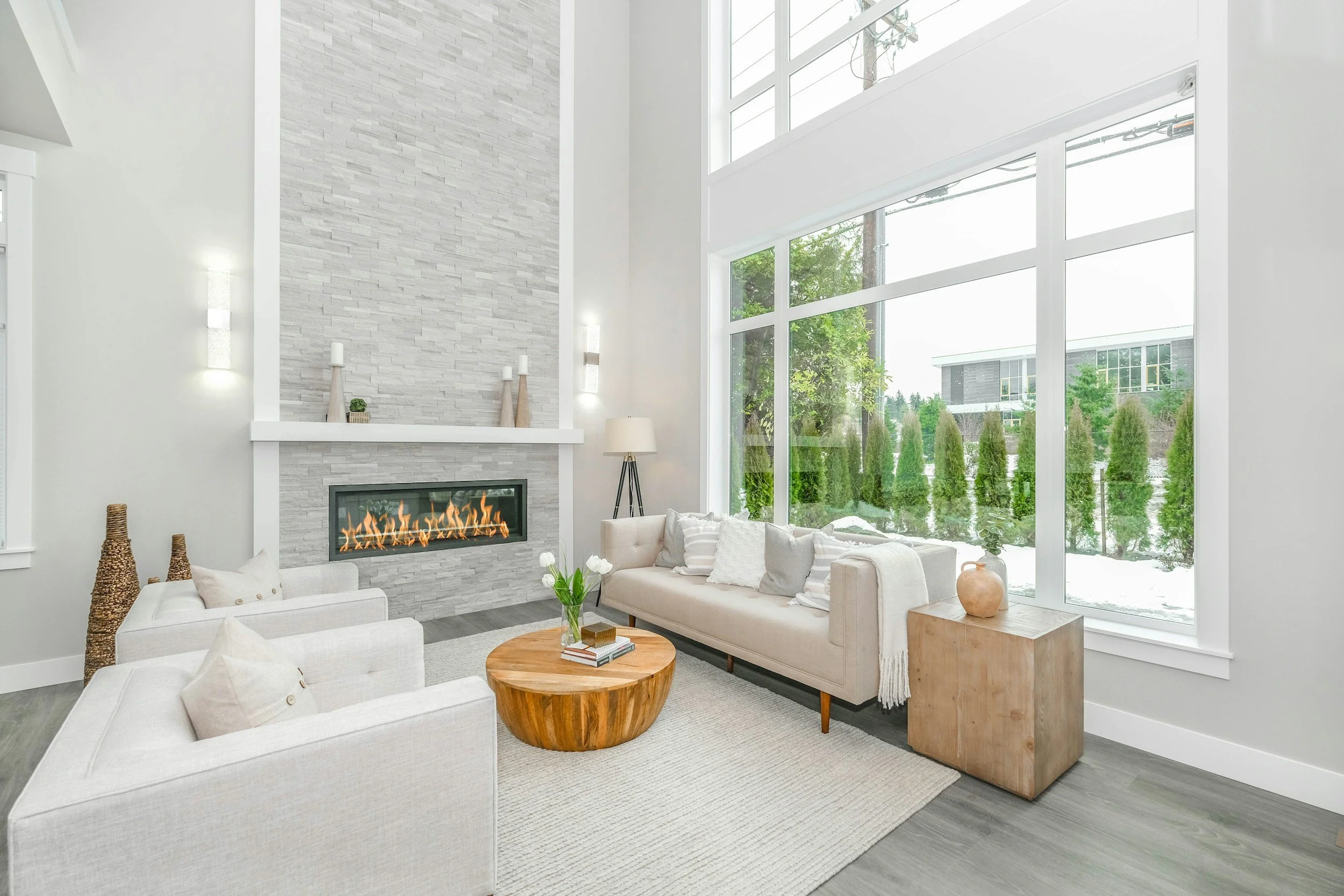 Modern living room with large windows, white sofas, a stone fireplace, and decorative vases.