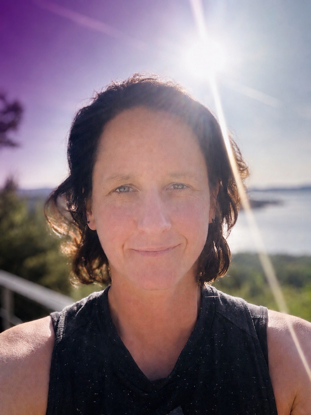 Nahaufnahme einer Frau mit dunklen, kurzen Haaren, die in der Sonne steht. Im Hintergrund Wasser und Natur, Sonnenstrahlen im blauen Himmel.