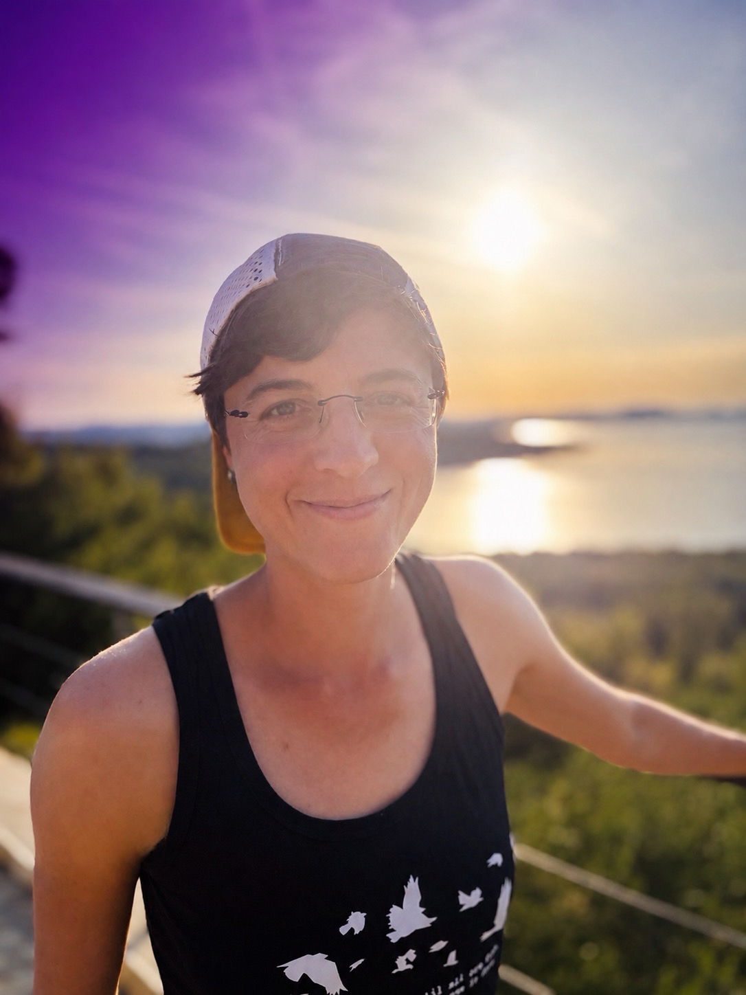 Frau lächelnd bei Sonnenuntergang am Wasser, trägt Sonnenbrille, schwarzen Tanktop mit Vogelmotiven, Cap rückwärts, im Freien.