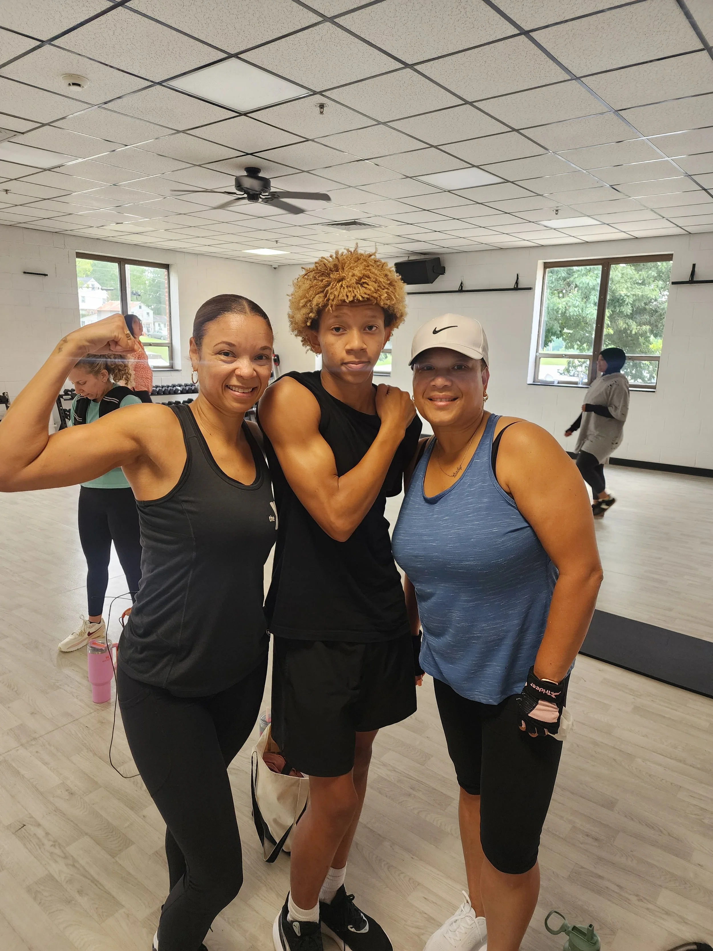 Three women posing together in a fitness studio, flexing arms and smiling. Two of them are wearing athletic tank tops, and the woman in the middle has curly blonde hair and is wearing a black tank top. The woman on the right wears a white cap. In the background, two other people are using the equipment and exercising.
