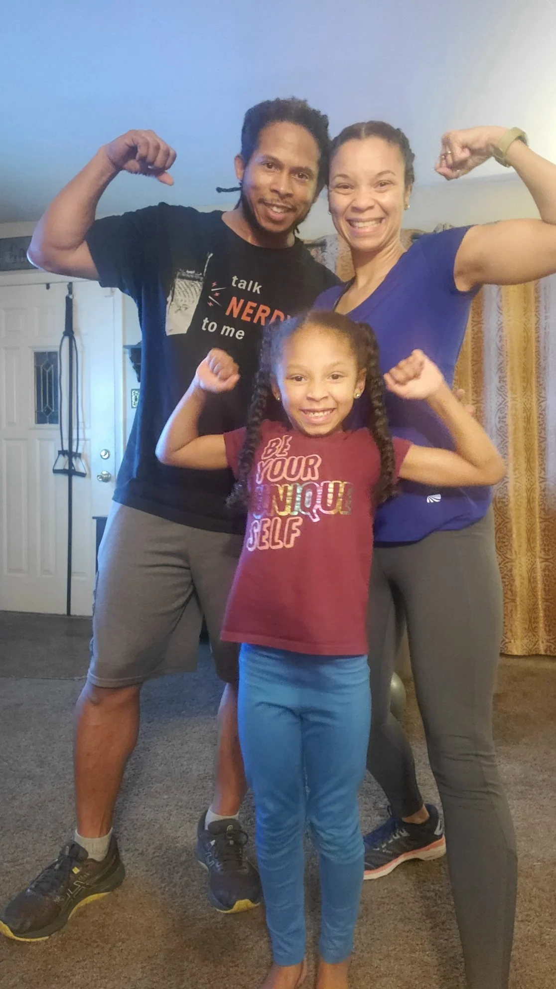 A family of three, including a man, woman, and young girl, are flexing their muscles and smiling at the camera inside a home. The man and woman are standing behind the girl, who is in the front.