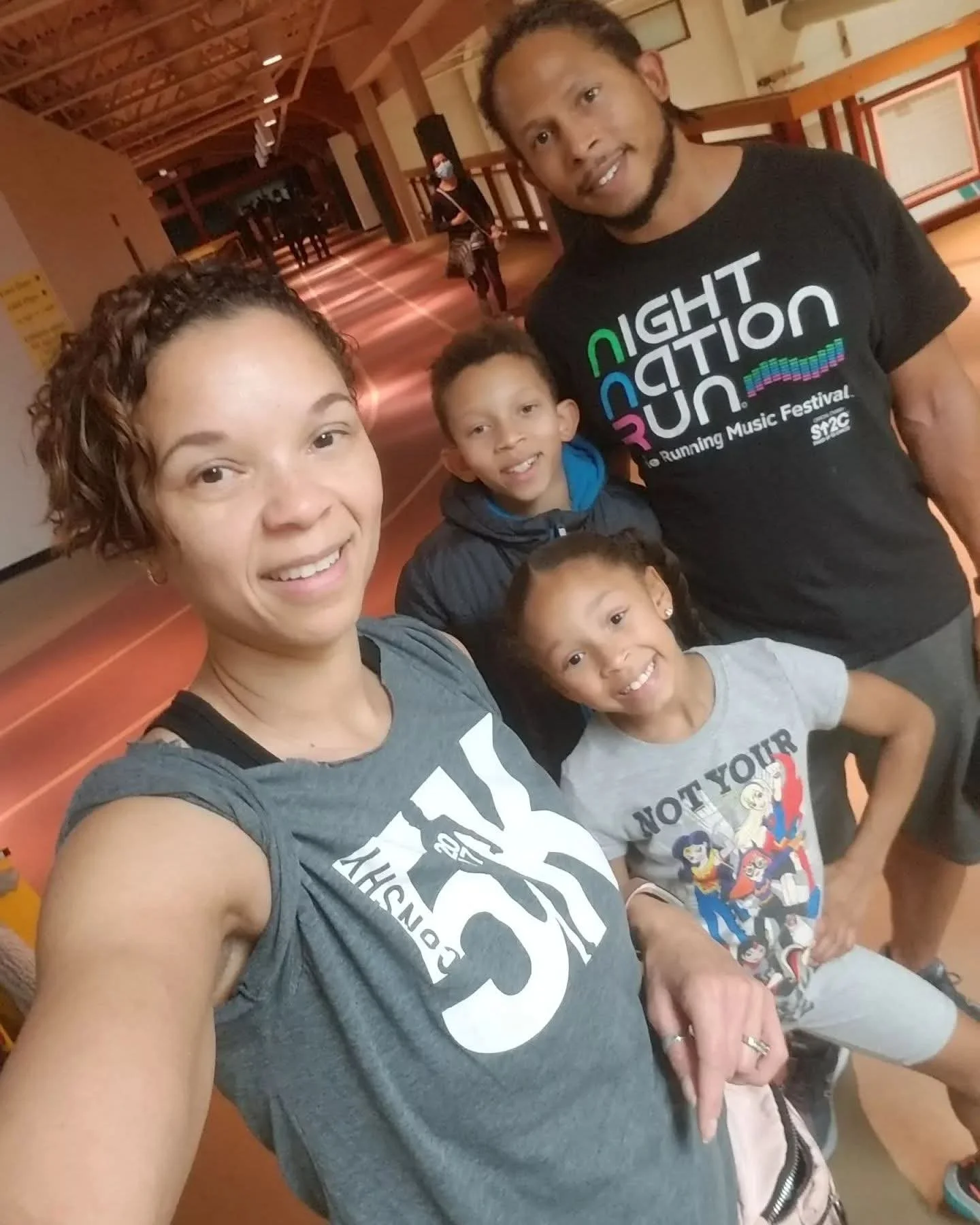 A group of four people taking a selfie in an indoor setting, with wooden ceiling beams. The woman in front has short curly hair, is smiling, and wears a gray T-shirt with black text. A young girl with dark curly hair and a gray T-shirt with Wonder Woman graphics is standing next to her. A man in the back has long hair in a braid and a beard, wearing a black T-shirt with colorful text that reads "Night Action Sun" and additional smaller text. Behind them, another woman wearing a face mask and casual clothing is in the background.