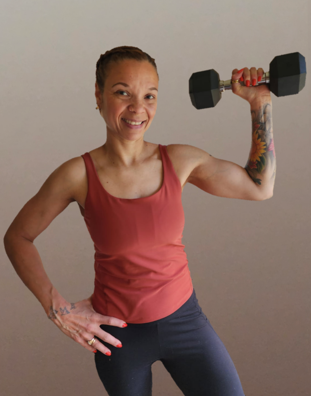 A woman with short hair, tattoos, and a red sleeveless top holding a black dumbbell in her right hand, smiling and standing against a plain background.