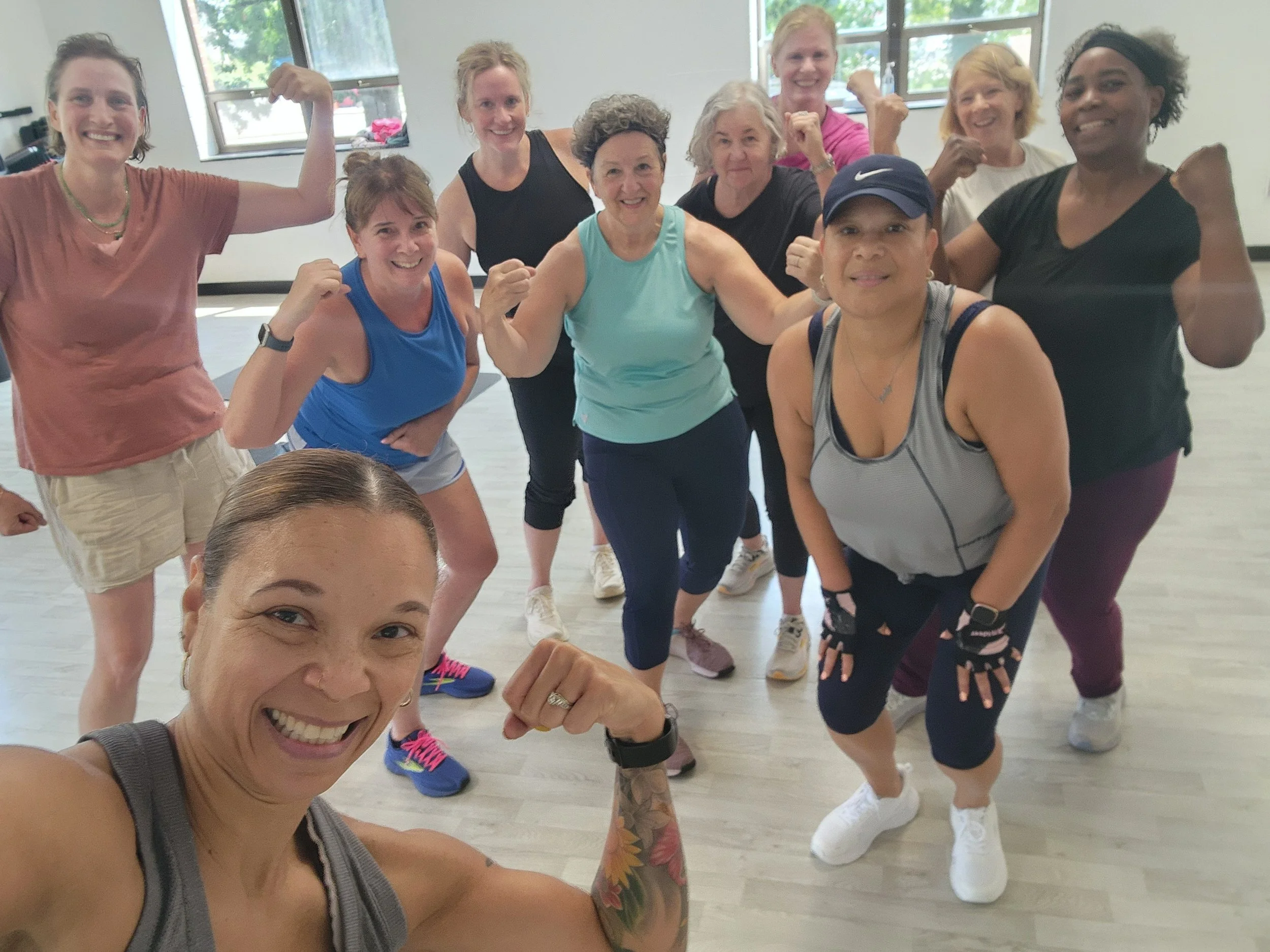 A group of ten women in workout attire posing in a fitness studio, flexing their muscles and smiling at the camera.