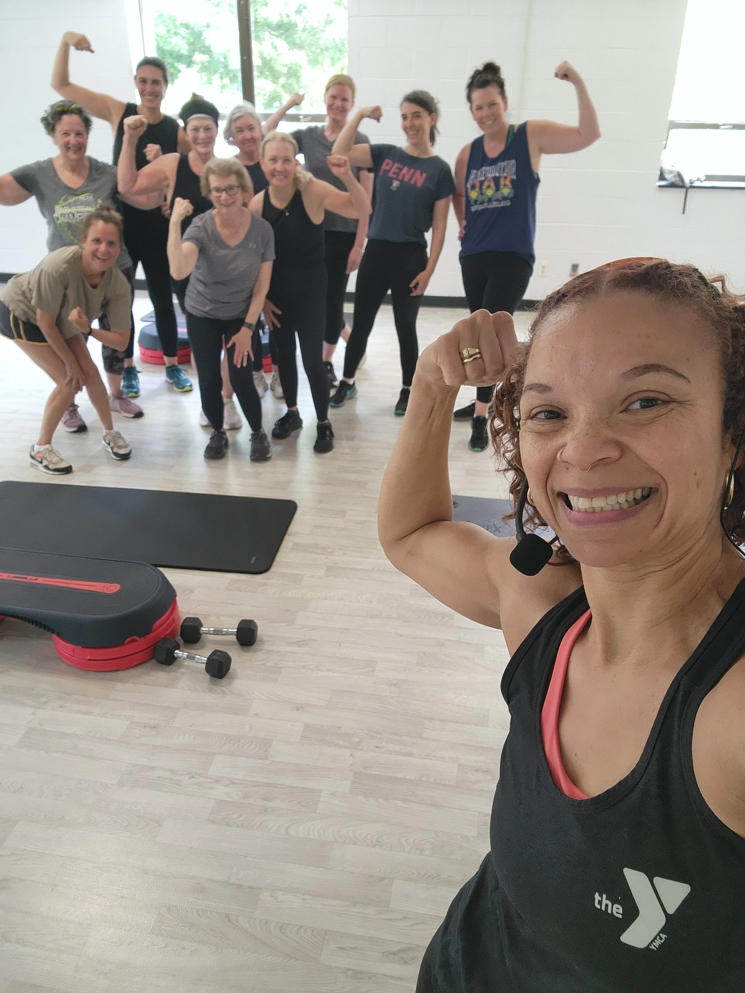 A group of women in workout attire flexing their biceps and smiling in a gym studio, with fitness equipment like dumbbells and a step platform visible on the floor. A woman in the foreground takes a selfie, showing her smiling face, curly hair, and a headset, wearing a black tank top with the YMCA logo.