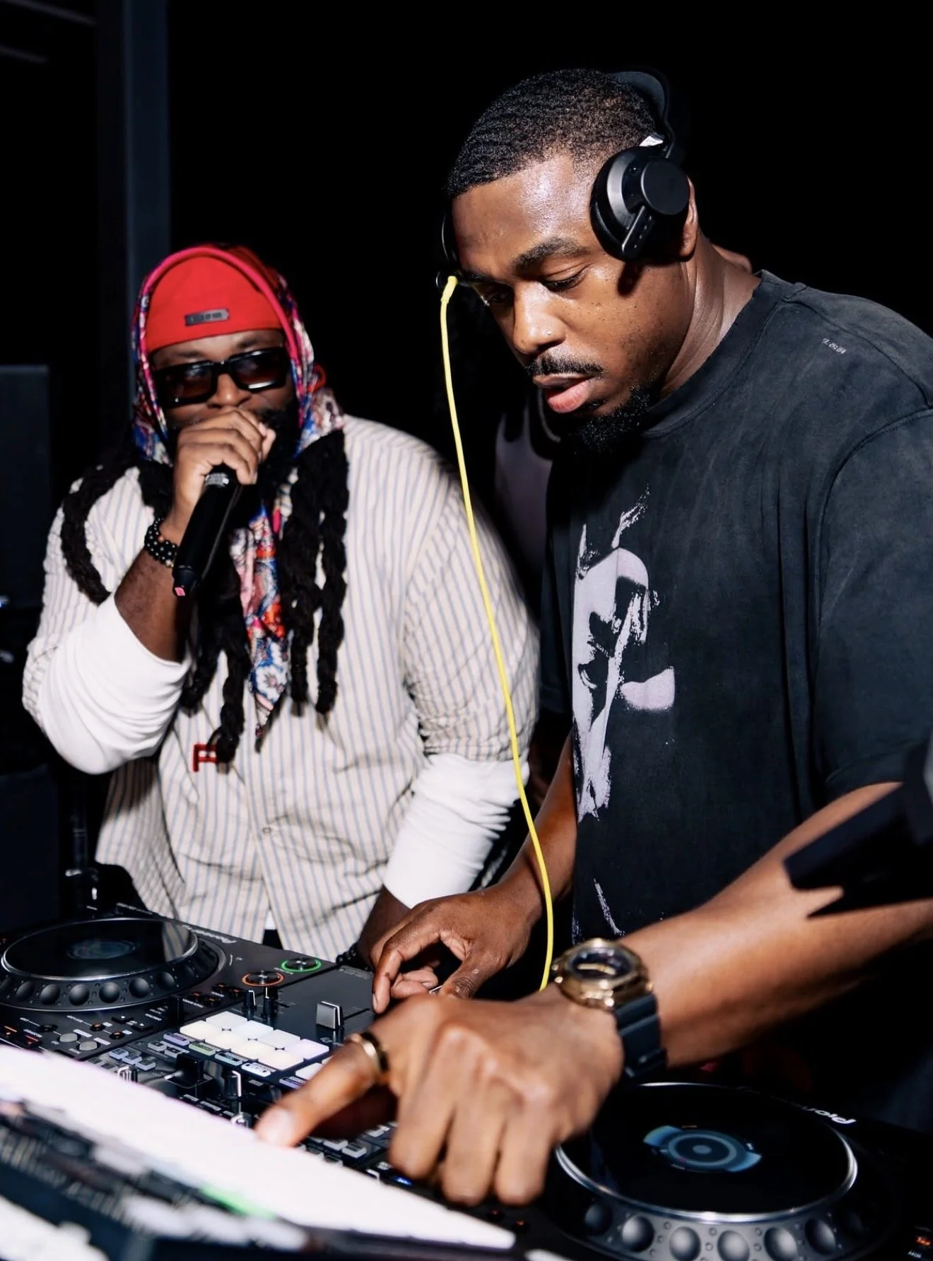 Two men at a DJ setup, with one wearing headphones and adjusting controls, and the other holding a microphone, both appearing focused during a music performance.