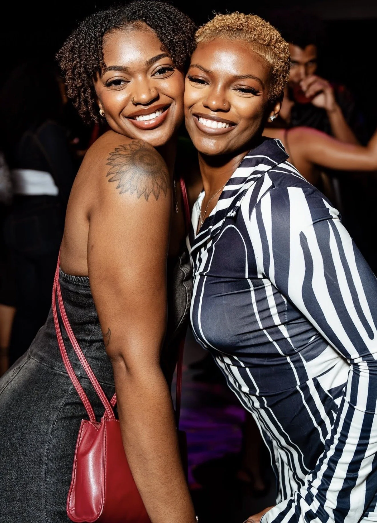 Two women smiling and hugging in a dark background, one with short, curly blonde hair and the other with medium-length, curly dark hair, both wearing stylish clothing and jewelry.