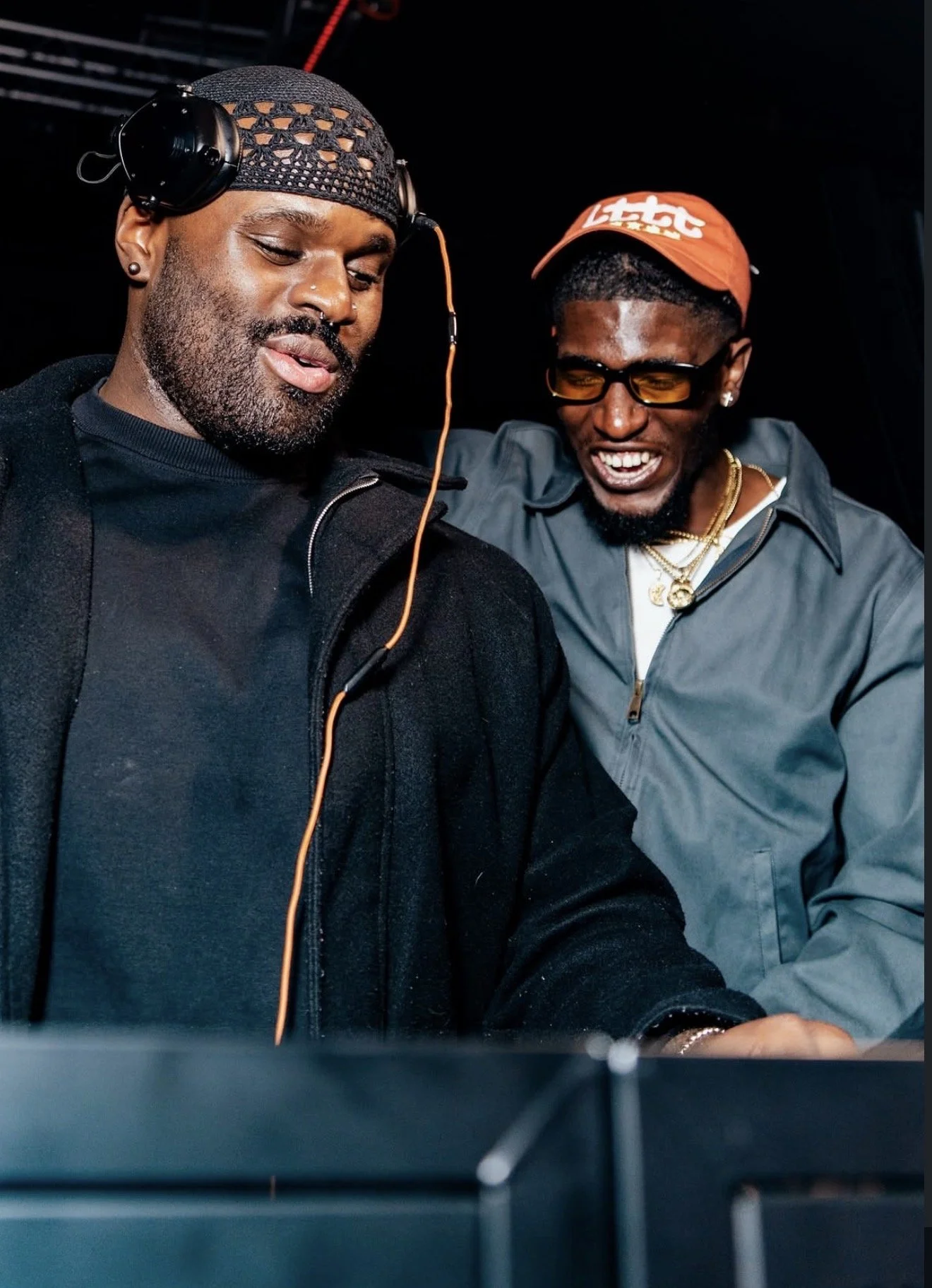 Two men smiling and enjoying themselves in a dark environment, possibly at a music event or club. The man on the left is wearing a black netted cap with headphones, and the man on the right is wearing glasses, a red cap, and jewelry.