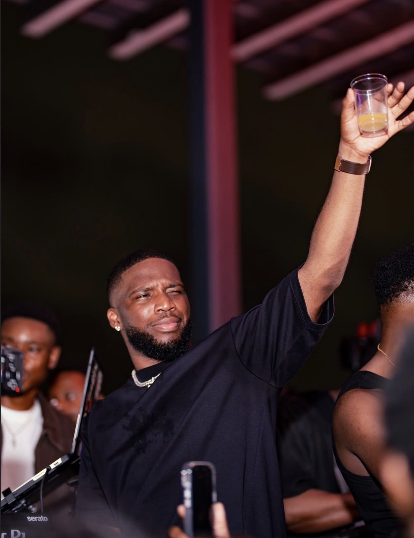 A man with a beard and earrings raising a glass in a dark setting with other people in the background.