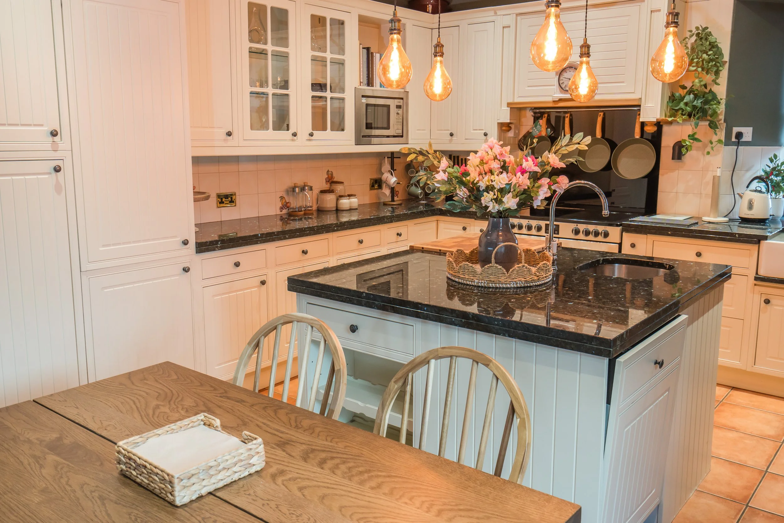 A cozy kitchen with cream-colored cabinets, black granite countertops, and a black stove. A bouquet of pink flowers is on the kitchen island, illuminated by hanging Edison bulbs. A wooden table with chairs is in the foreground.