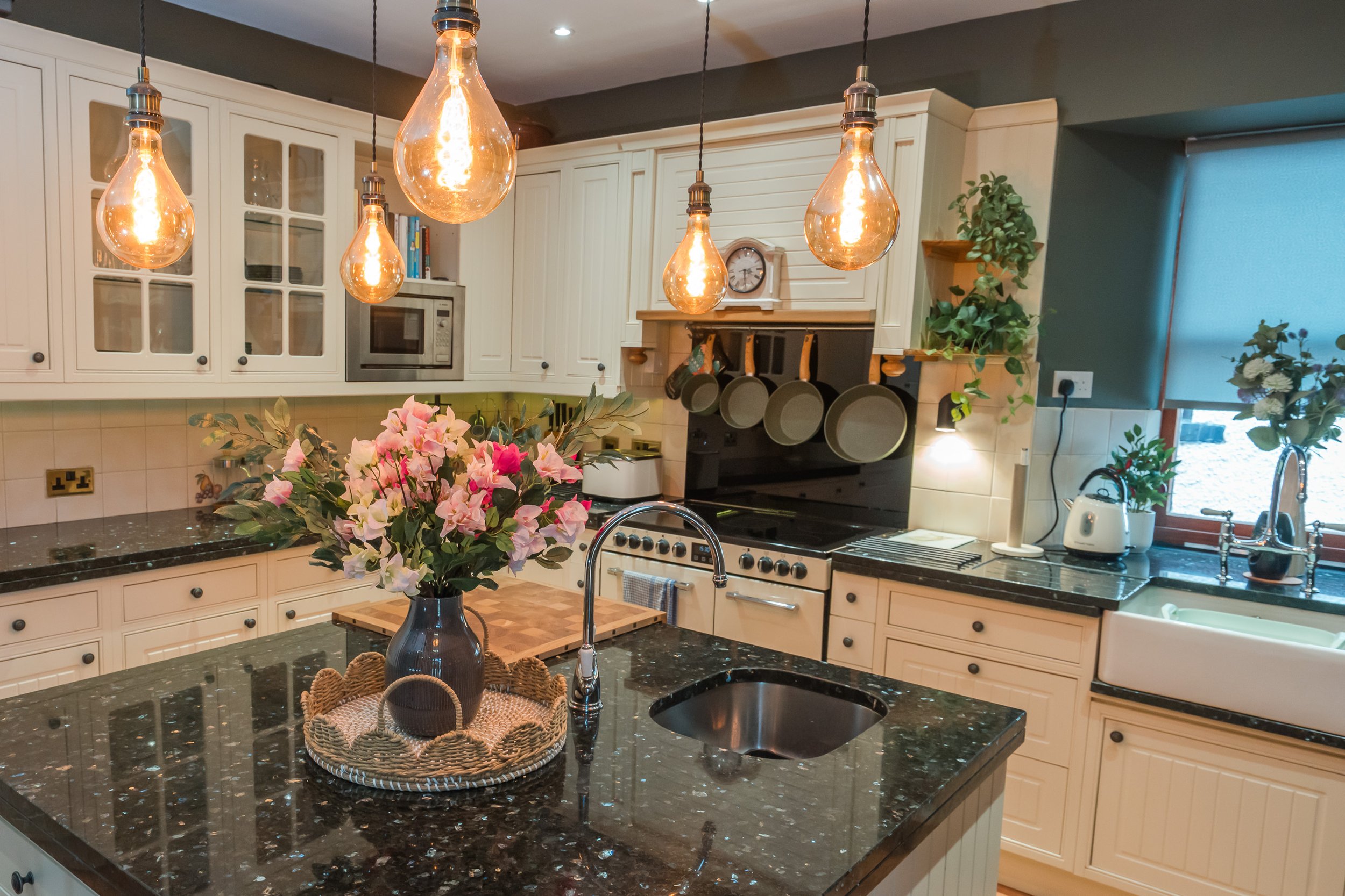 A modern kitchen with white cabinetry, black granite countertops, and a black stove. There are five hanging Edison-style light bulbs above the kitchen island, which has a bouquet of pink and cream flowers in a dark vase. The kitchen also features a m