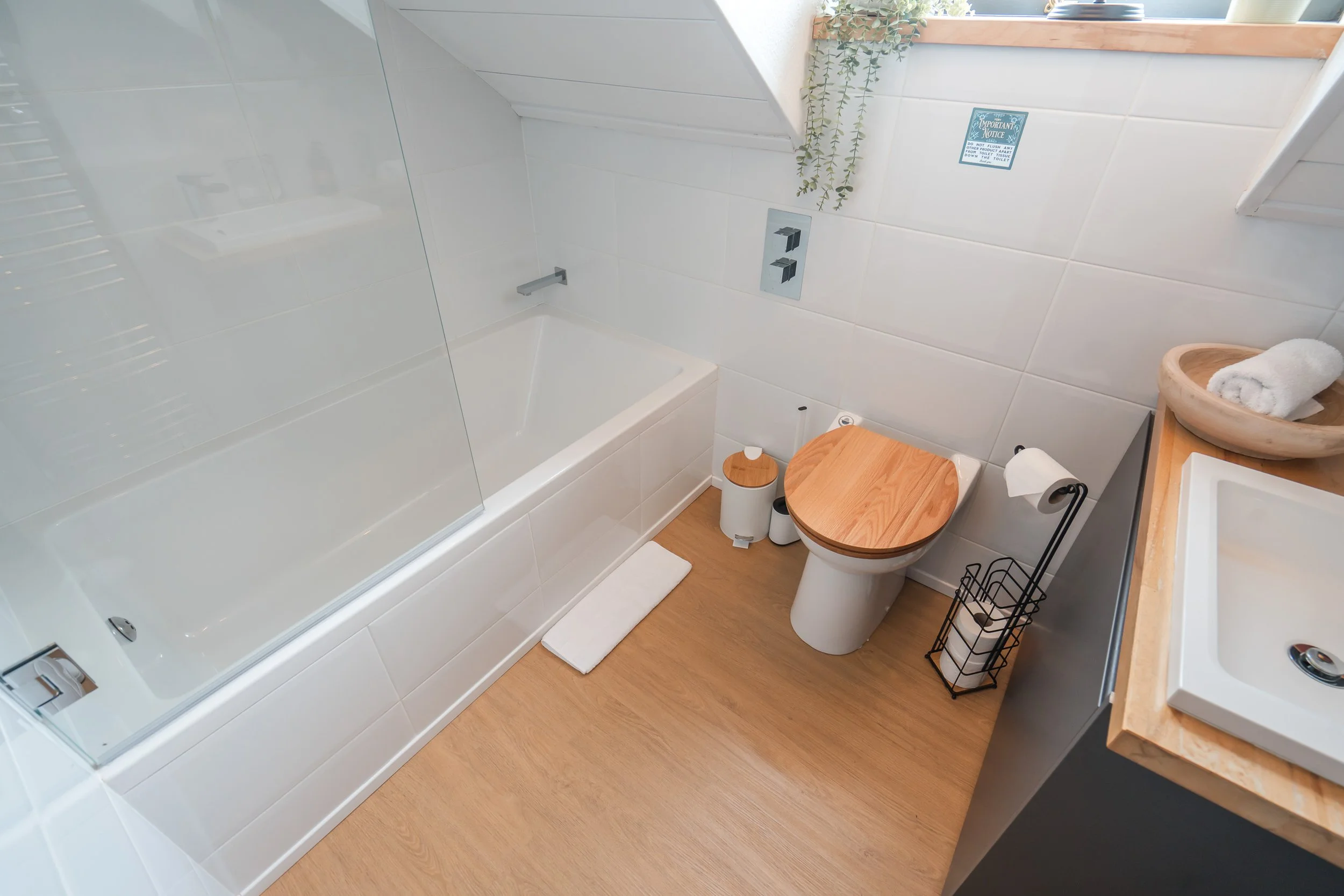 A modern bathroom with a white bathtub, a wooden toilet seat, a small white trash can, a bath mat, and a window with plants on the sill. There is also a sign on the wall near the window.