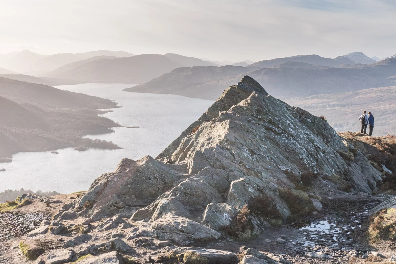 Two hikers standing on Ben A'an overlooking a lake and mountain range.