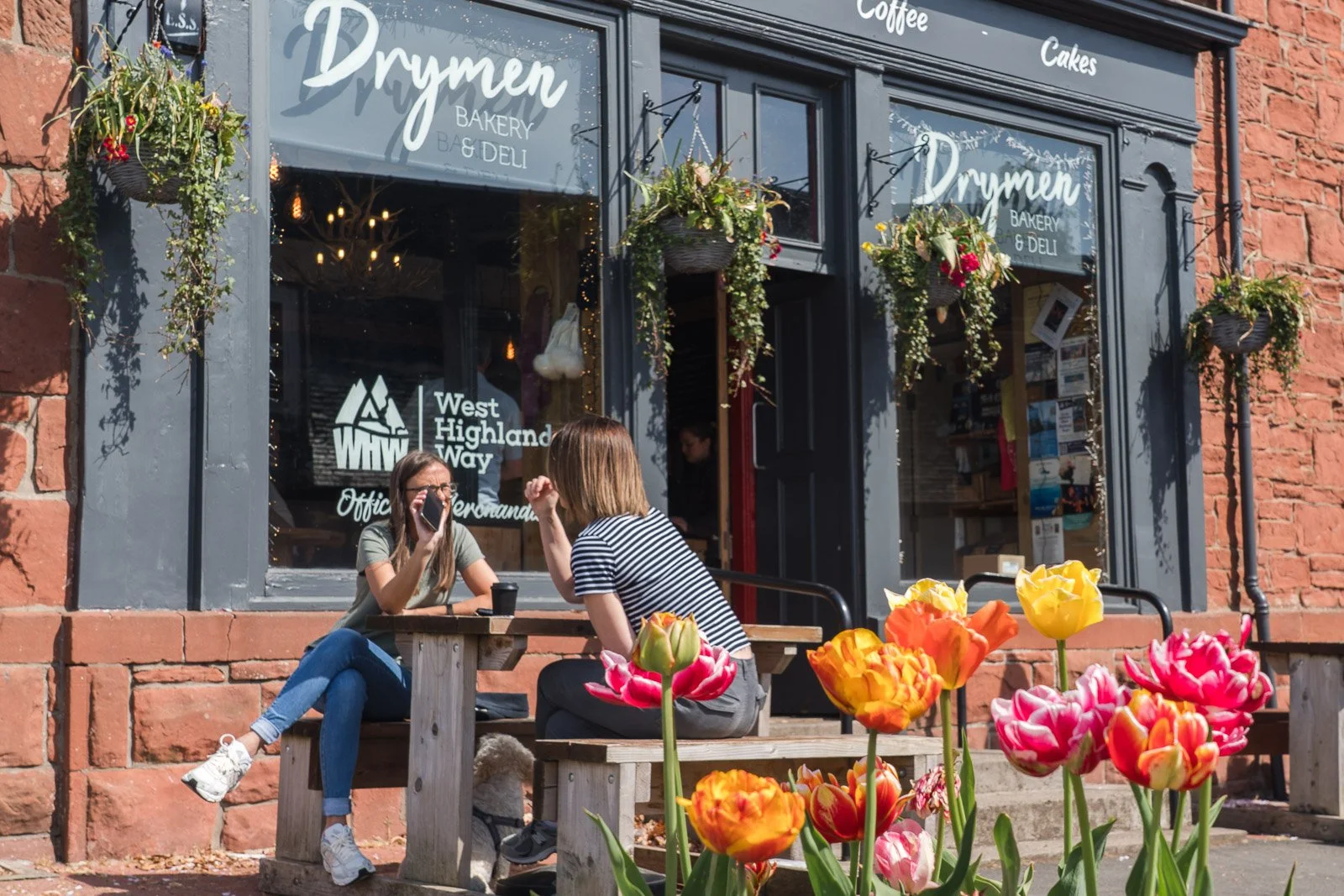 Two women sit at a wooden picnic table outside a bakery and deli, engaged in conversation. Behind them, the bakery's window displays the name 'Drymen Bakery & Deli' and the logo of West Highland Way. In front of the table, colorful tulips bloom, adding vibrant color to the scene.