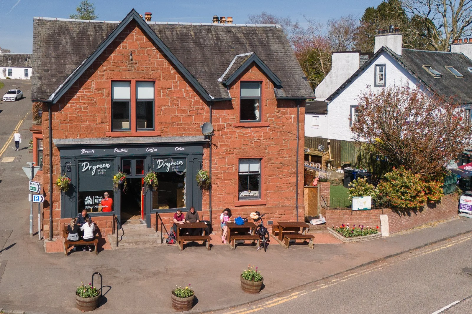 A two-story red brick bakery cafe named 'Drymen' with outdoor seating on a sidewalk; people are sitting at picnic tables in front of the shop, and there are hanging flower baskets on the storefront.