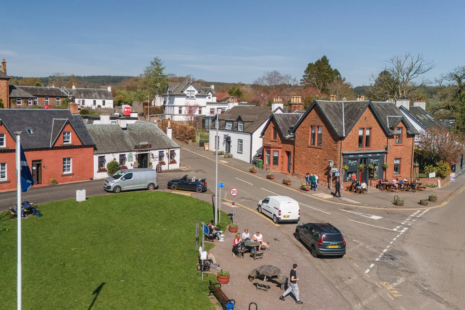 Drymen village scene on a sunny day with a grassy area with benches and people sitting, a street with cars parked, and old brick and white houses, some with shops, on a corner. Trees with leaves and a hill in the background.