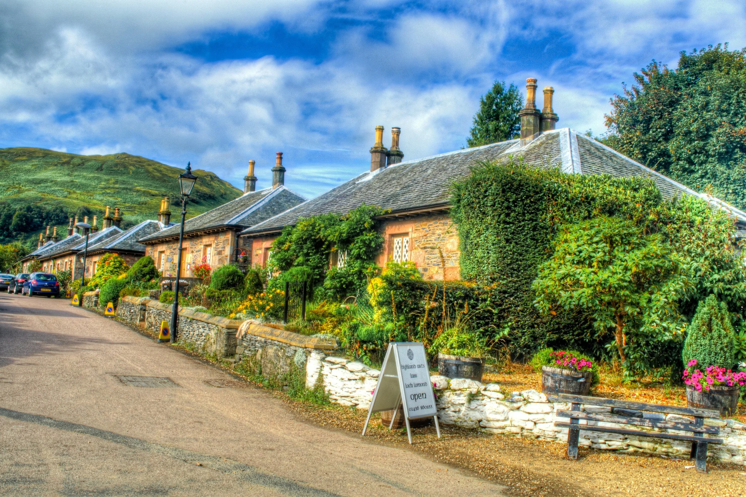 a row of stone cottages in Luss with chimneys, decorated with flowers and greenery, under a partly cloudy blue sky.