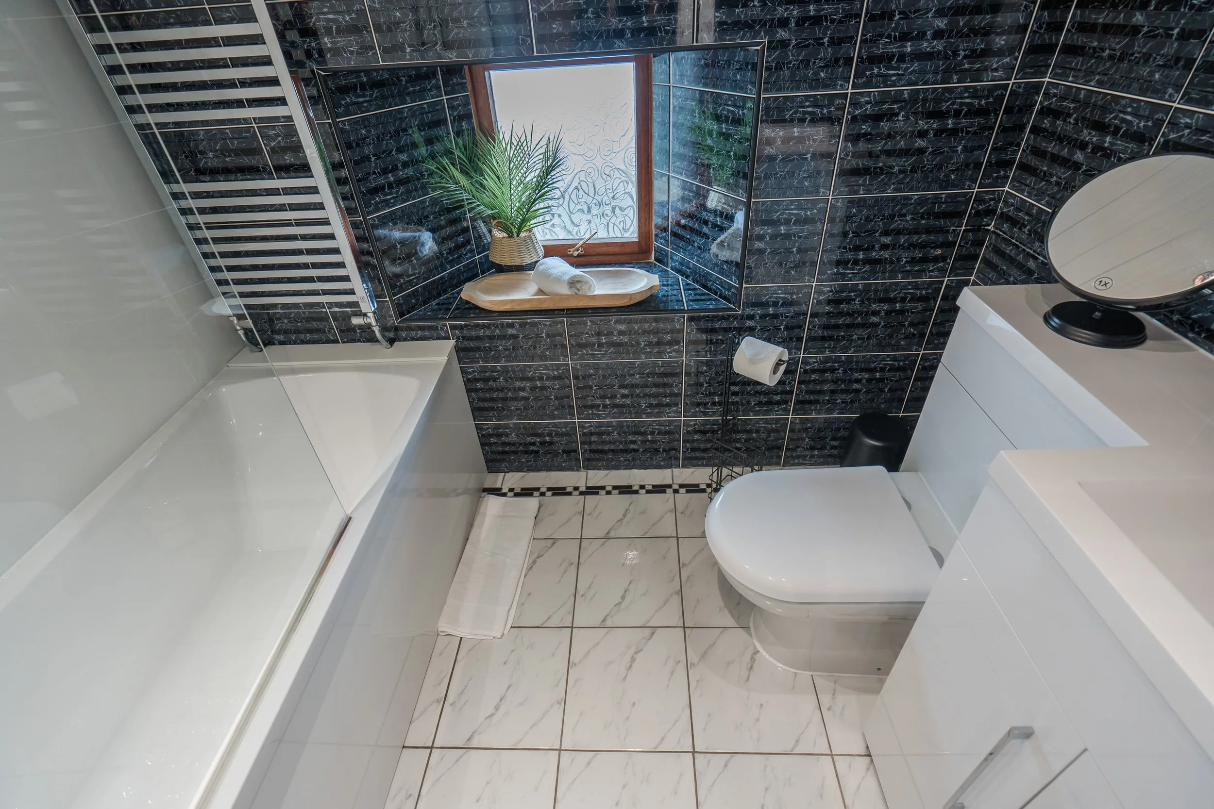 A modern bathroom featuring black marble tiles with gold accents on the walls, a white toilet, a glass shower door, a window with a potted plant and towel, and a small vanity with a mirror.