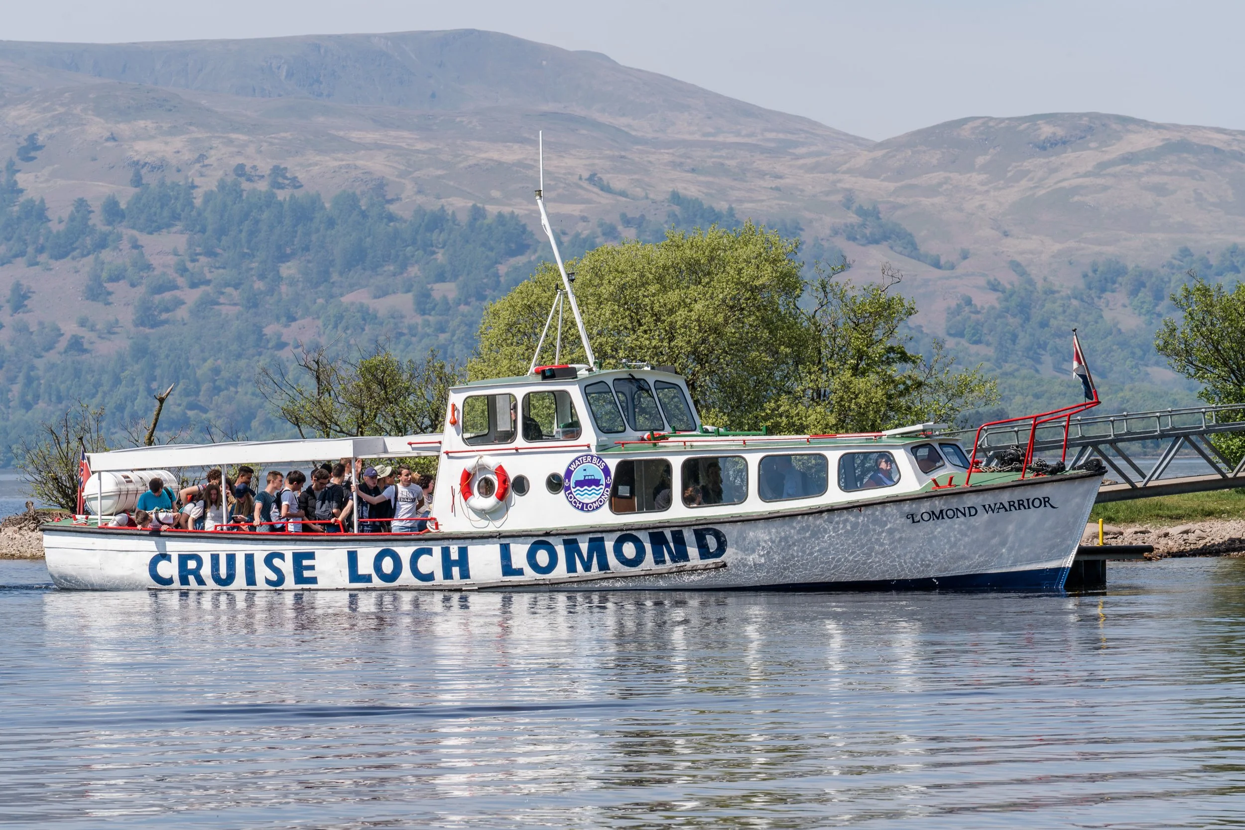 Cruise Loch Lomond boat named 'Lomond Warrior' with people on board, cruising on a lake surrounded by green trees and hills in the background.