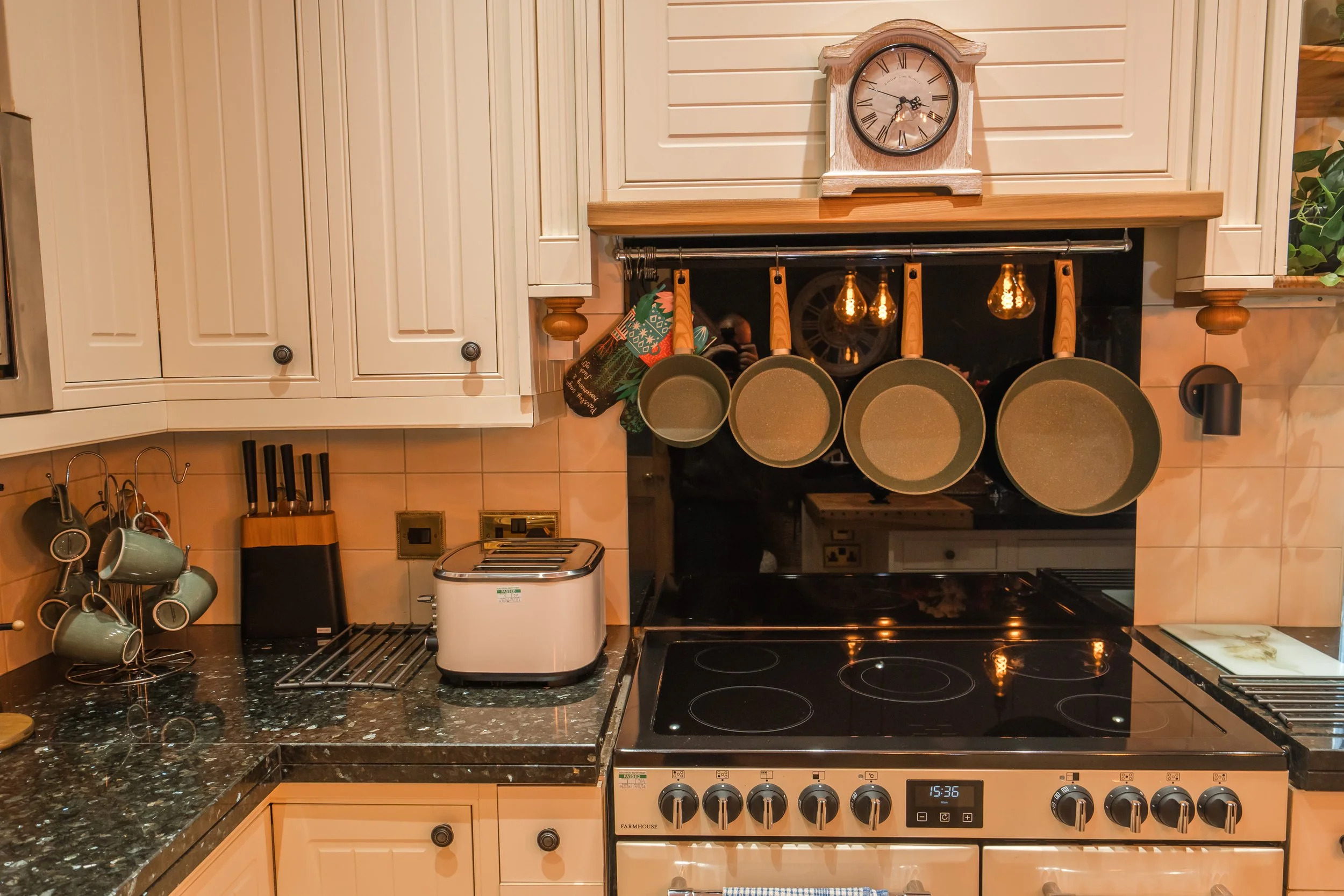 Kitchen stove with four burners, black stovetop, and digital clock showing 15:36. Above it, hanging from a wooden rail, are four frying pans with wooden handles, and a clock on the wall. To the left, there are some coffee cups hanging on hooks and a 
