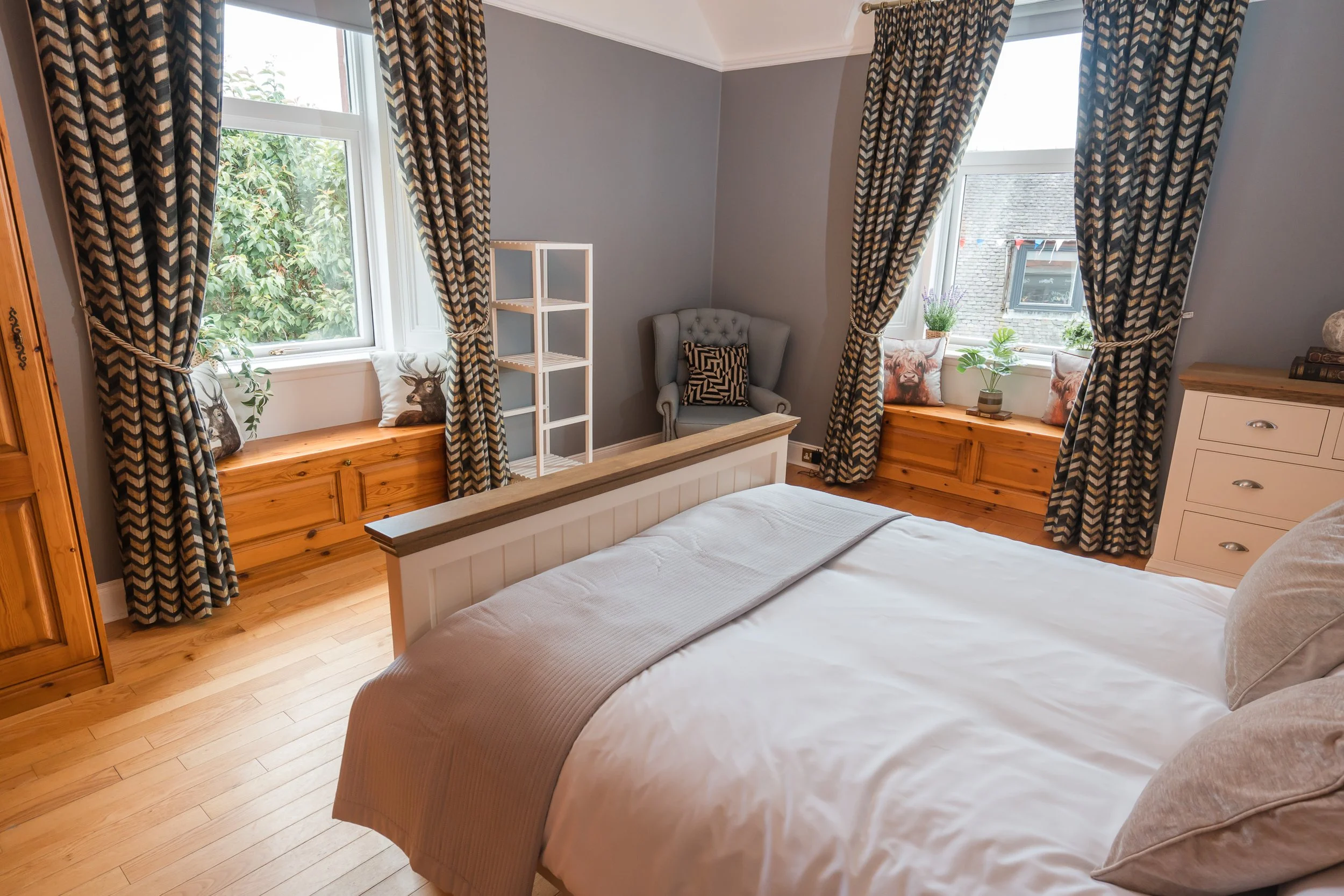 A cozy bedroom with a bed in the foreground, featuring a beige bedspread and pillows. There are three windows with patterned curtains pulled back, revealing outdoor greenery and buildings. The windows have wooden window seats with decorative pillows,
