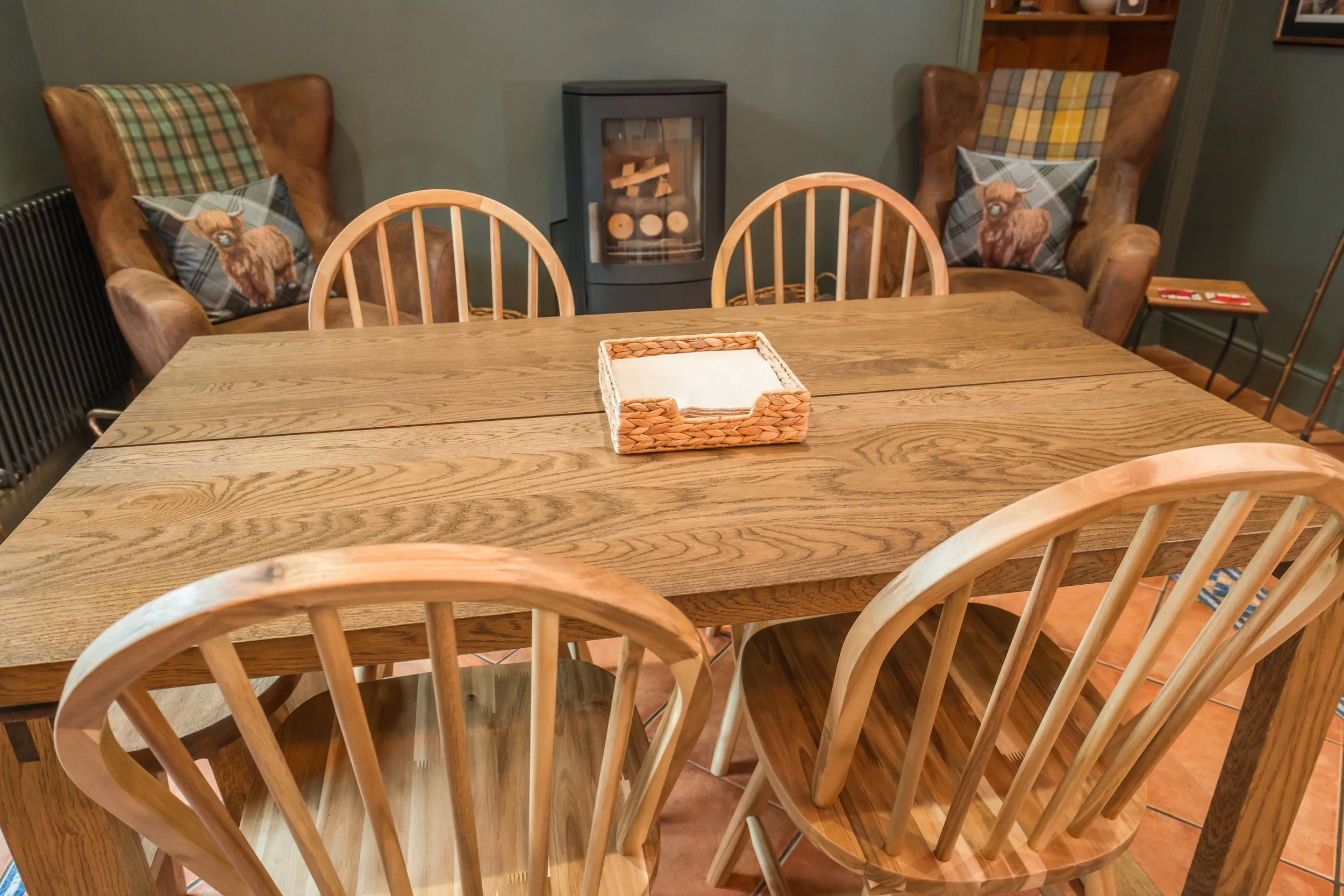 A wooden dining table with six matching chairs in a room with green walls and brown tiled flooring. Two wingback chairs with plaid cushions and llama-themed pillows are positioned against the wall, with a wood stove and a small side table in the back
