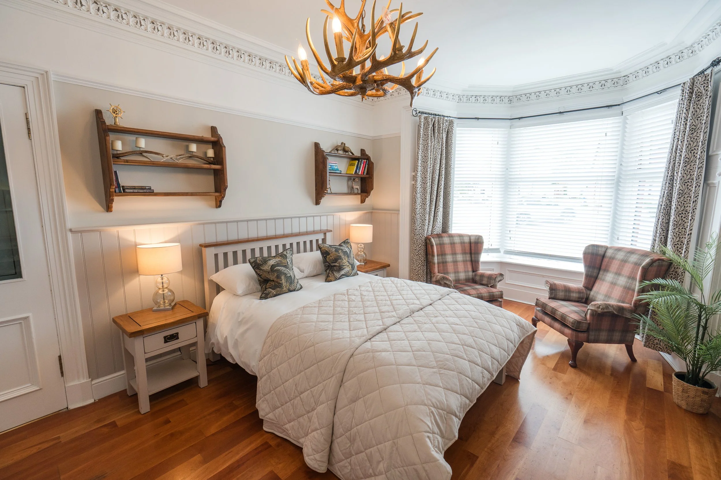 A bedroom with a large bay window with blinds and patterned curtains, a bed with white linens and decorative pillows, wooden bedside tables with lamps, and a plaid armchair near a potted plant, with a chandelier made of antlers hanging from the ceili