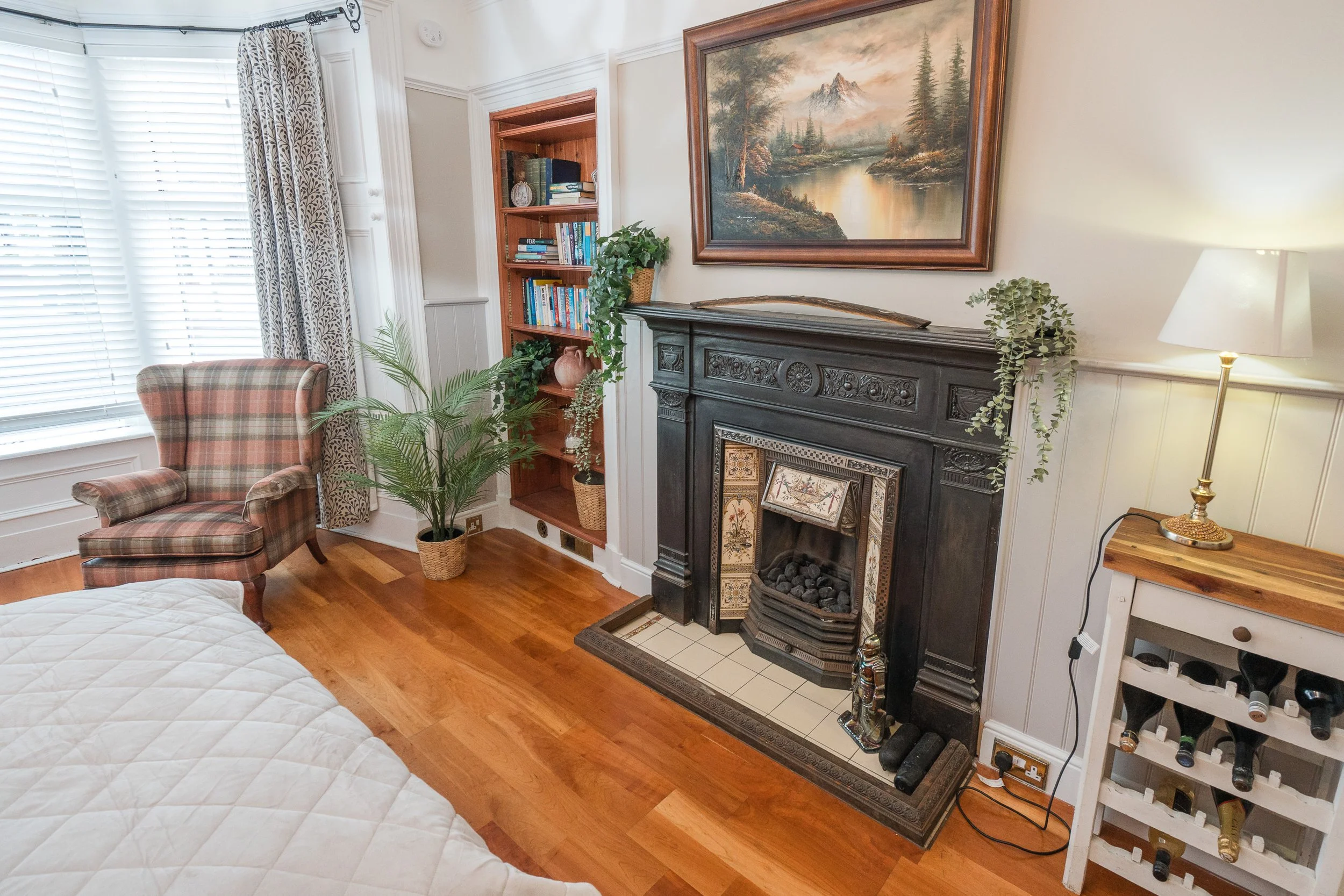 Cozy living room corner with a plaid armchair, a tall wooden bookshelf filled with books and plants, a black fireplace with decorative tiles, a landscape painting above the fireplace, a table with a lamp, and hardwood floors with white walls.