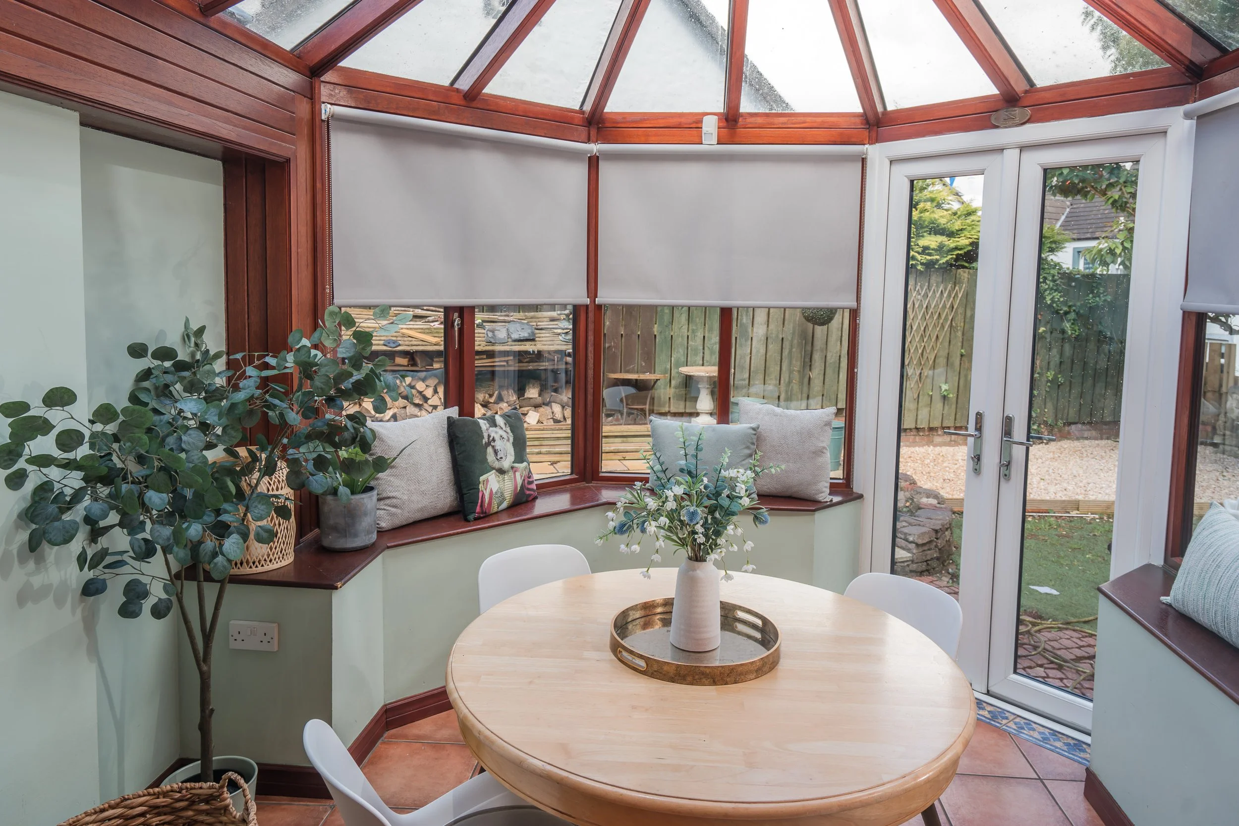 A conservatory with curved glass roof and windows, a round wooden table with a white vase containing flowers, and a green wall with a large potted eucalyptus tree. There's a corner window seat with cushions, and a view of the garden outside.