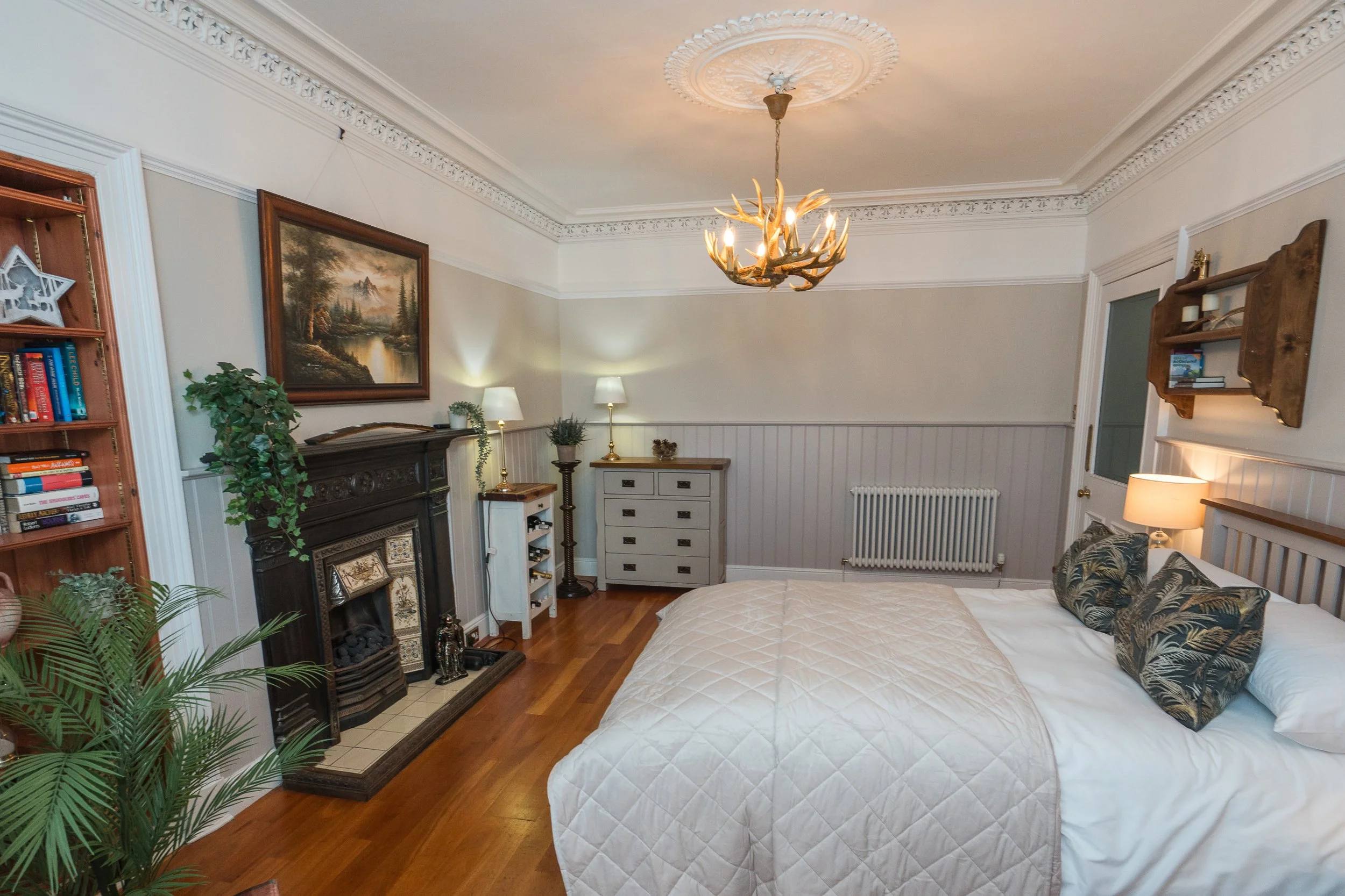 A cosy Drymen Holiday bedroom with a fireplace, nightstand, lamp, and a large bed with tropical print pillows, decorated with a chandelier and artwork of nature on the walls.