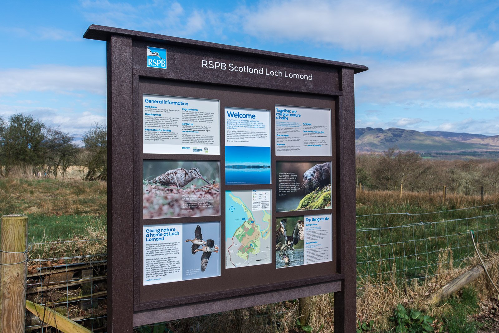 Information board at RSPB Scotland Loch Lomond showing details about wildlife, nature conservation, and visitor information, with background of grassy landscape and hills under a partly cloudy sky.