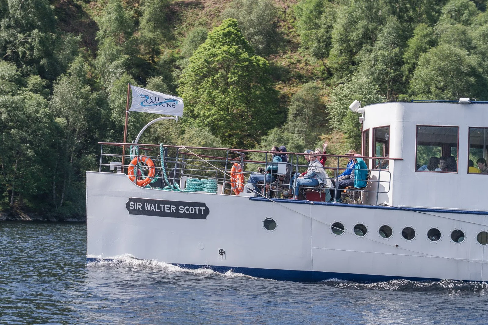 Steamship Sir Walter Scott sailing on a river with a group of people on deck, some waving, surrounded by trees and greenery in the background.