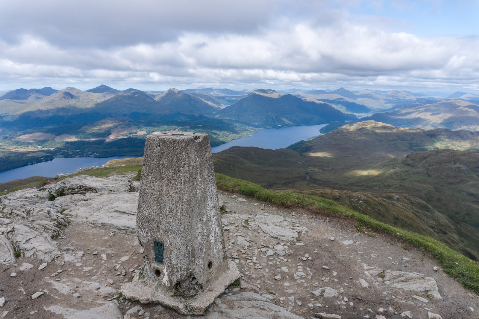 View from a Ben Lomond with a stone trig point overlooking lakes and rolling hills under a cloudy sky.