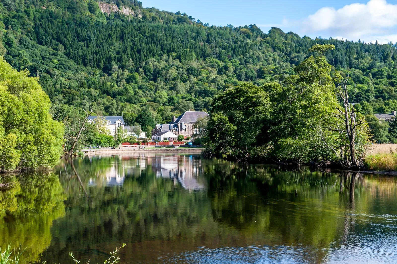 Callander river scene with green trees along the riverbank, reflections in the water, and houses in the background at the base of a forested hill under a partly cloudy sky.