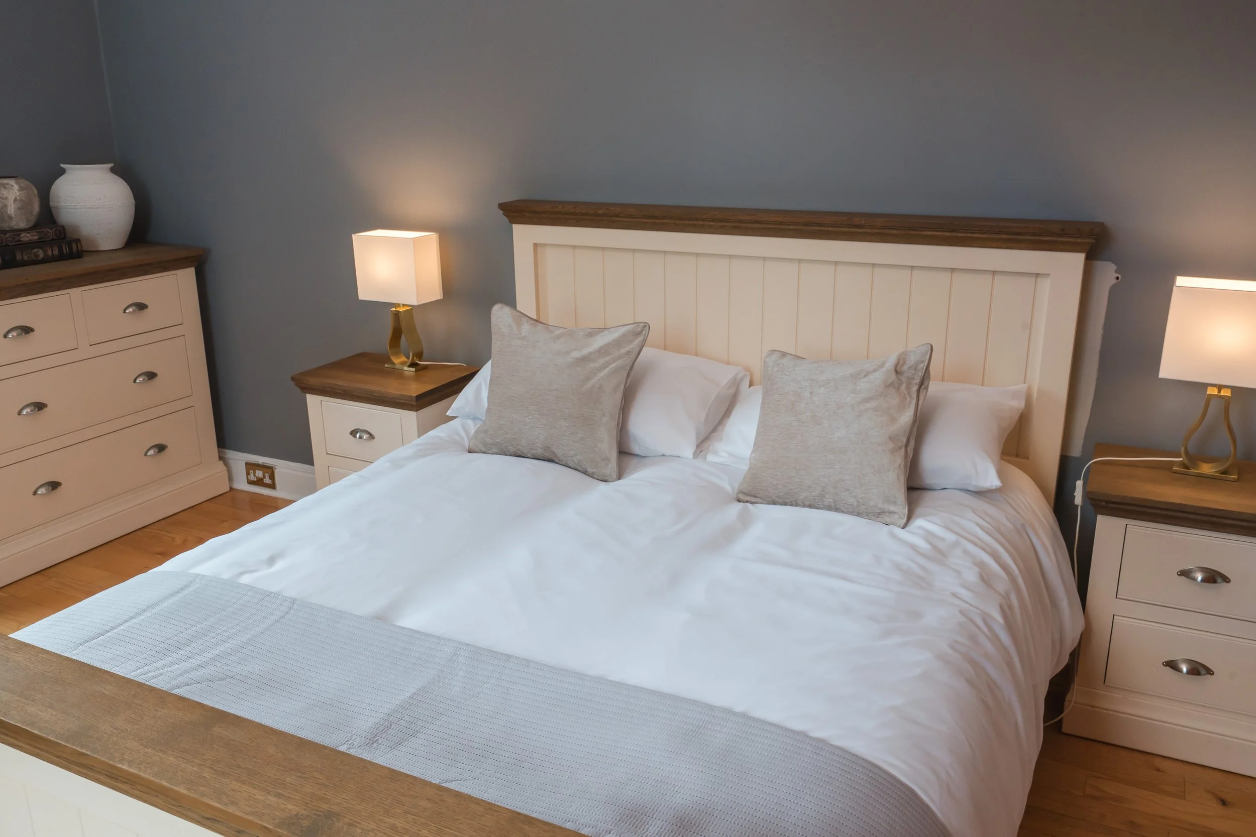A bedroom with a white wooden bed frame and beige bedding, accented with beige pillows. Two matching nightstands with lamps are on either side of the bed. A beige dresser with decorative items is on the left. The walls are painted blue, and hardwood 