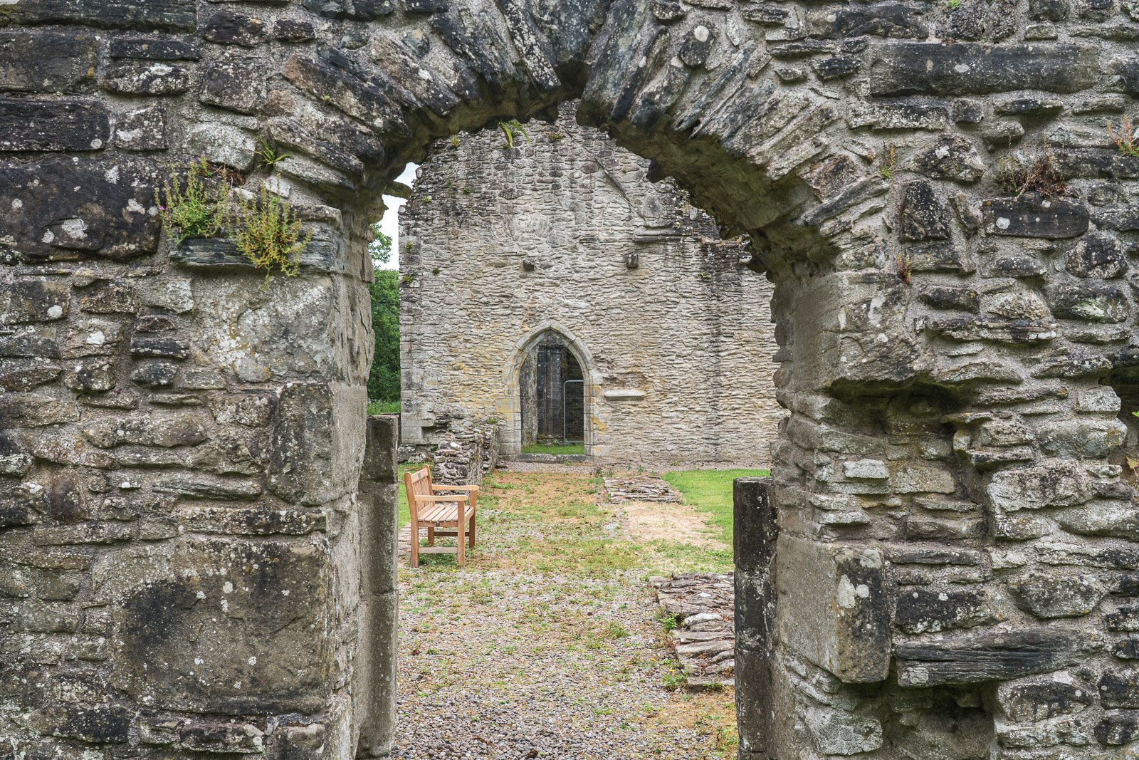 Inchmahome Priory with t stone archway looking into a historic church ruins with a wooden bench and grassy pathway.