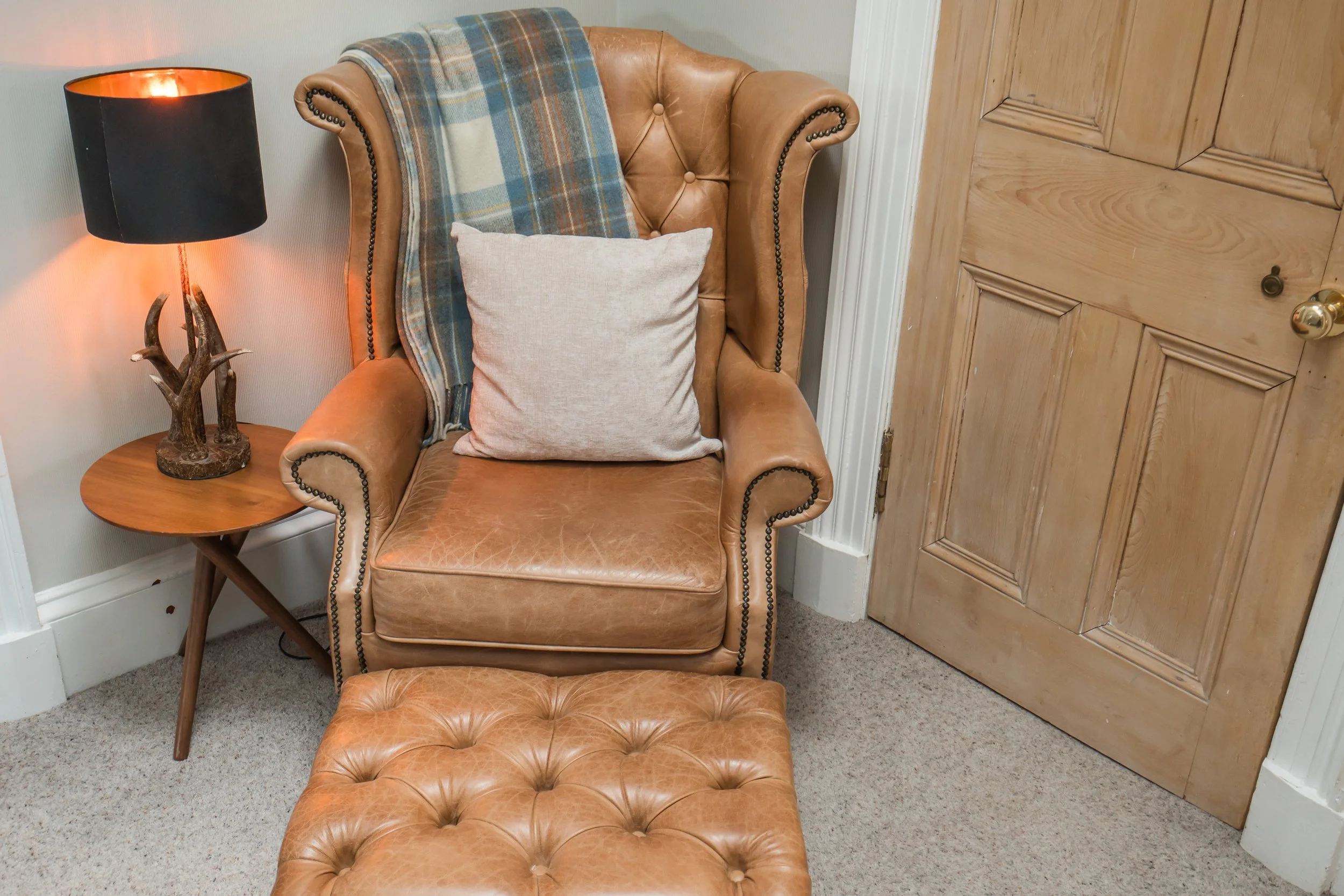 A leather armchair with nailhead trim, a blanket draped over the back, and a cushion on the seat, beside a side table with a decorative antler lamp, next to a wooden door in a room with beige carpet and cream walls.