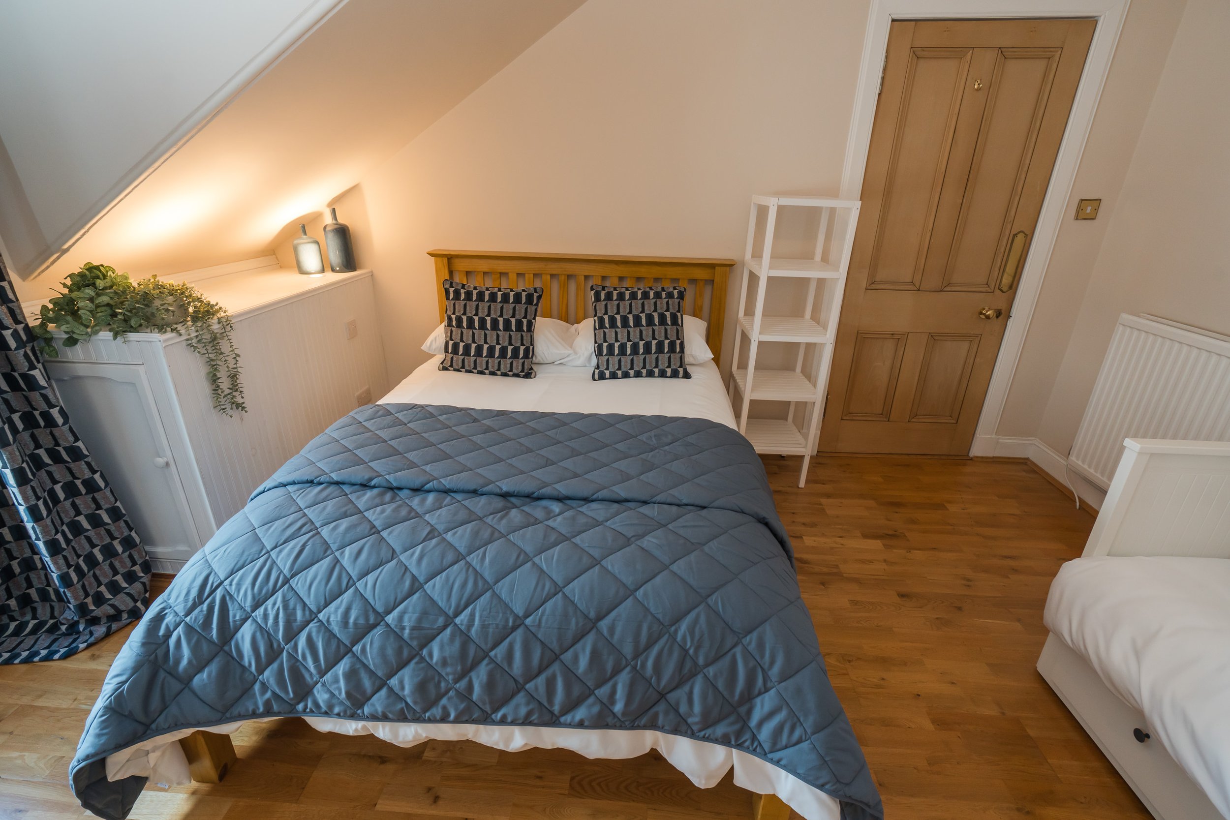 A cozy bedroom with a bed covered in a blue quilt, black and beige patterned pillows, and a wooden headboard. To the right, there is a white shelving unit next to a closed wooden door. On the left, a ledge with two vases and green plants. The room ha
