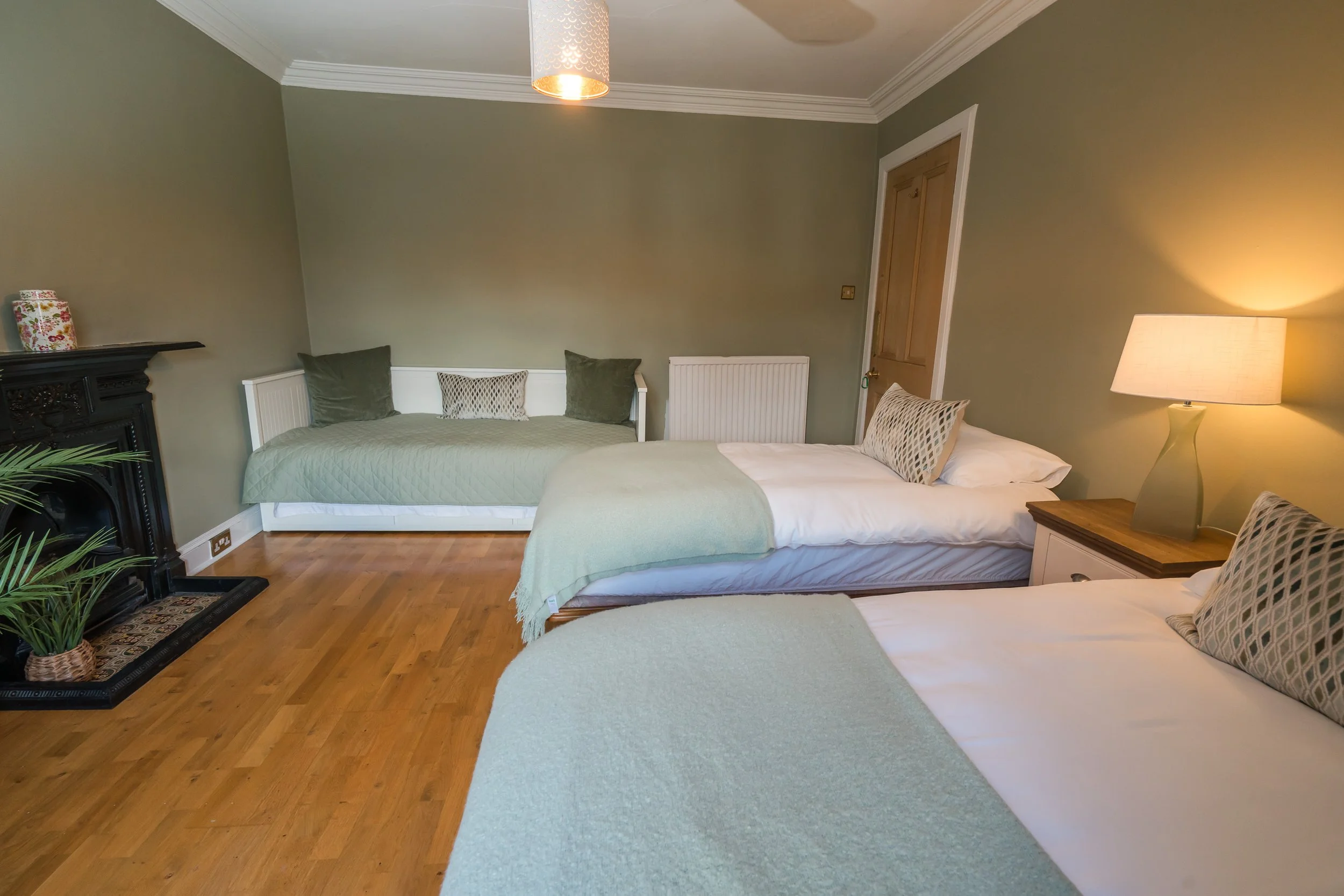 A room with three beds, green walls, wooden flooring, a black fireplace, and a wall-mounted light fixture.