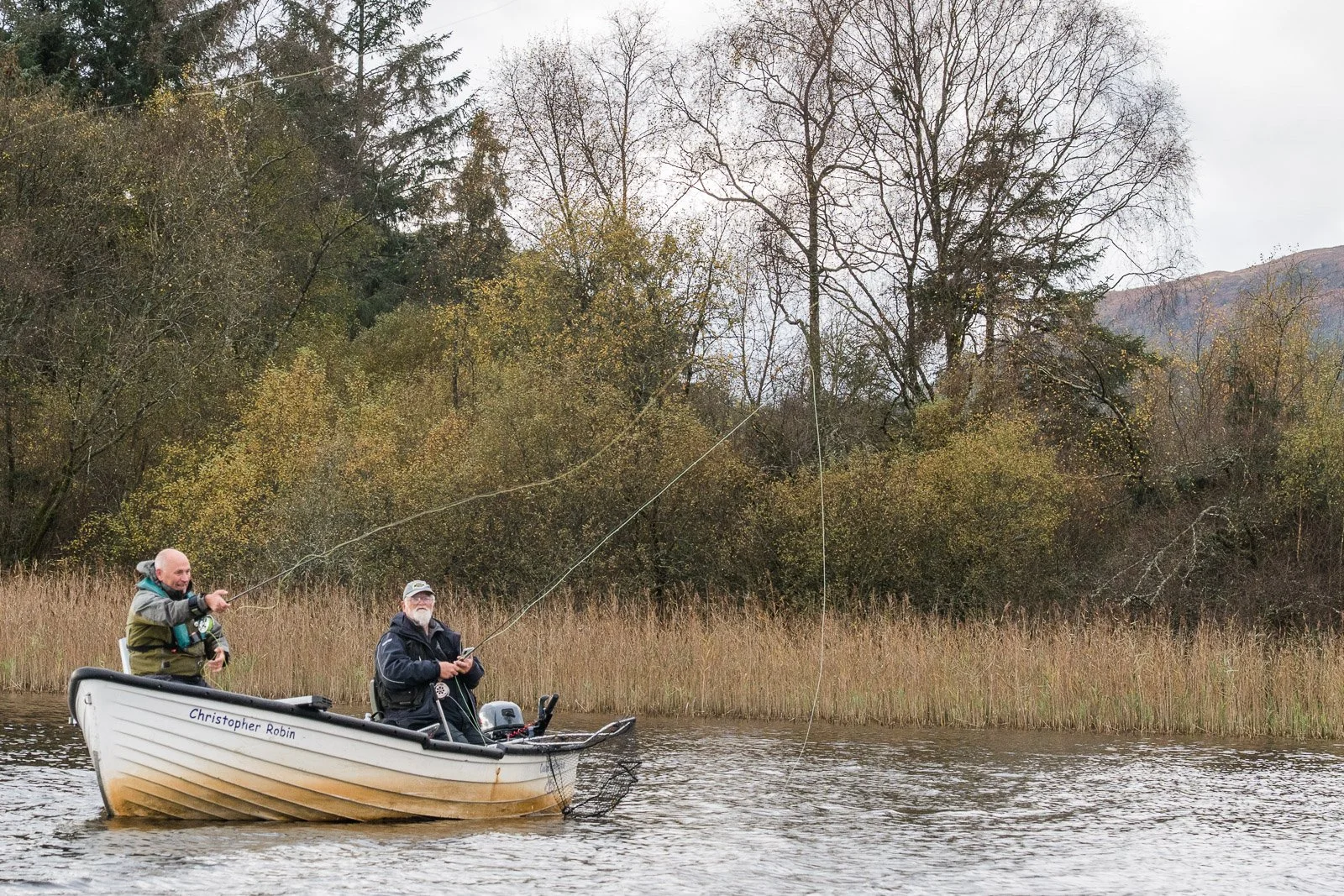 Two men fishing from a small boat named Christopher Robin on a Lake of Menteith, with trees and a mountain in the background on a cloudy day.