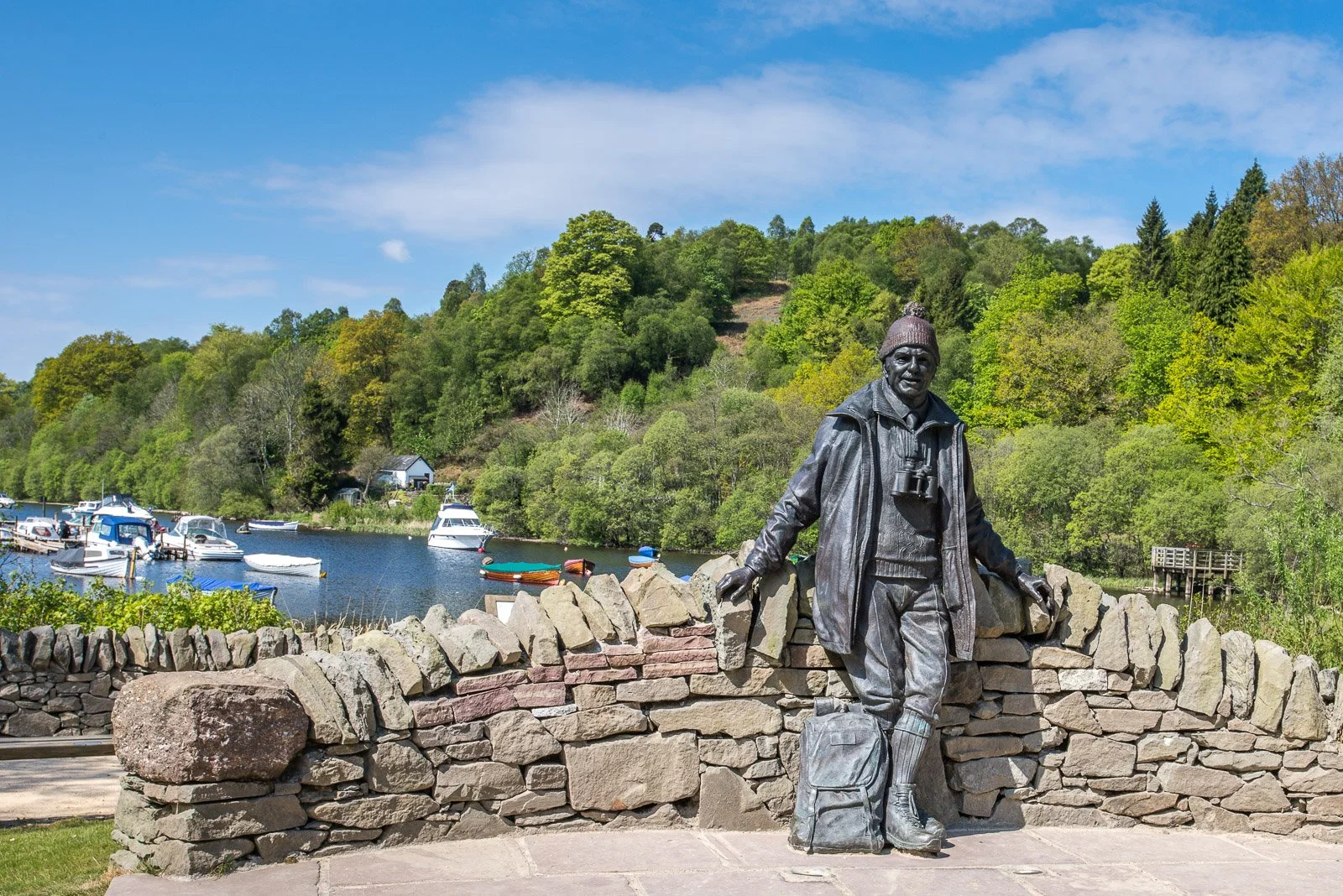 Tom Weir Statue at Balmaha, Loch Lomond outdoors near a river with boats, surrounded by trees and a stone wall.