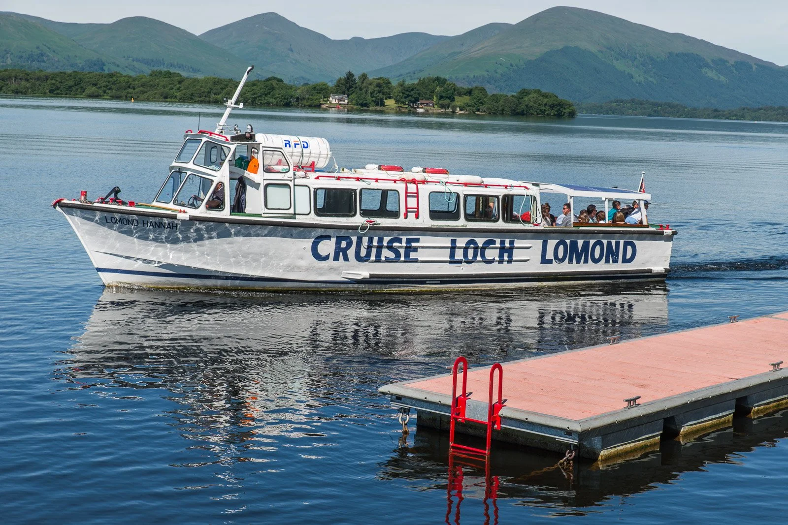 A white boat labeled 'CRUISE LOCH LOMOND' is sailing on a calm lake with mountains and greenery in the background. Passengers are on board, some sitting and some standing. A red ladder is attached to the dock in the foreground.