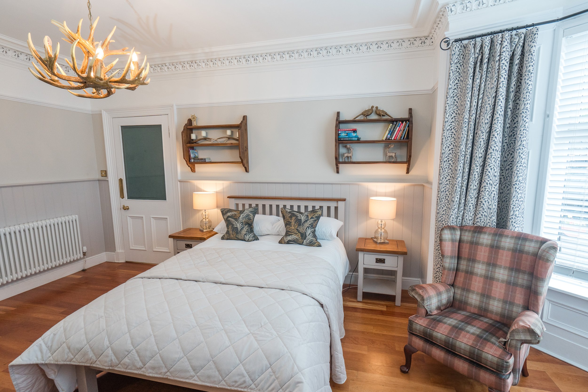 A cozy bedroom with two beds, wooden nightstands, and matching lamps. The bed has decorative pillows with leaf patterns. There are two small wooden shelves on the wall with books and decorative bird figurines. A plaid armchair is near a large window 