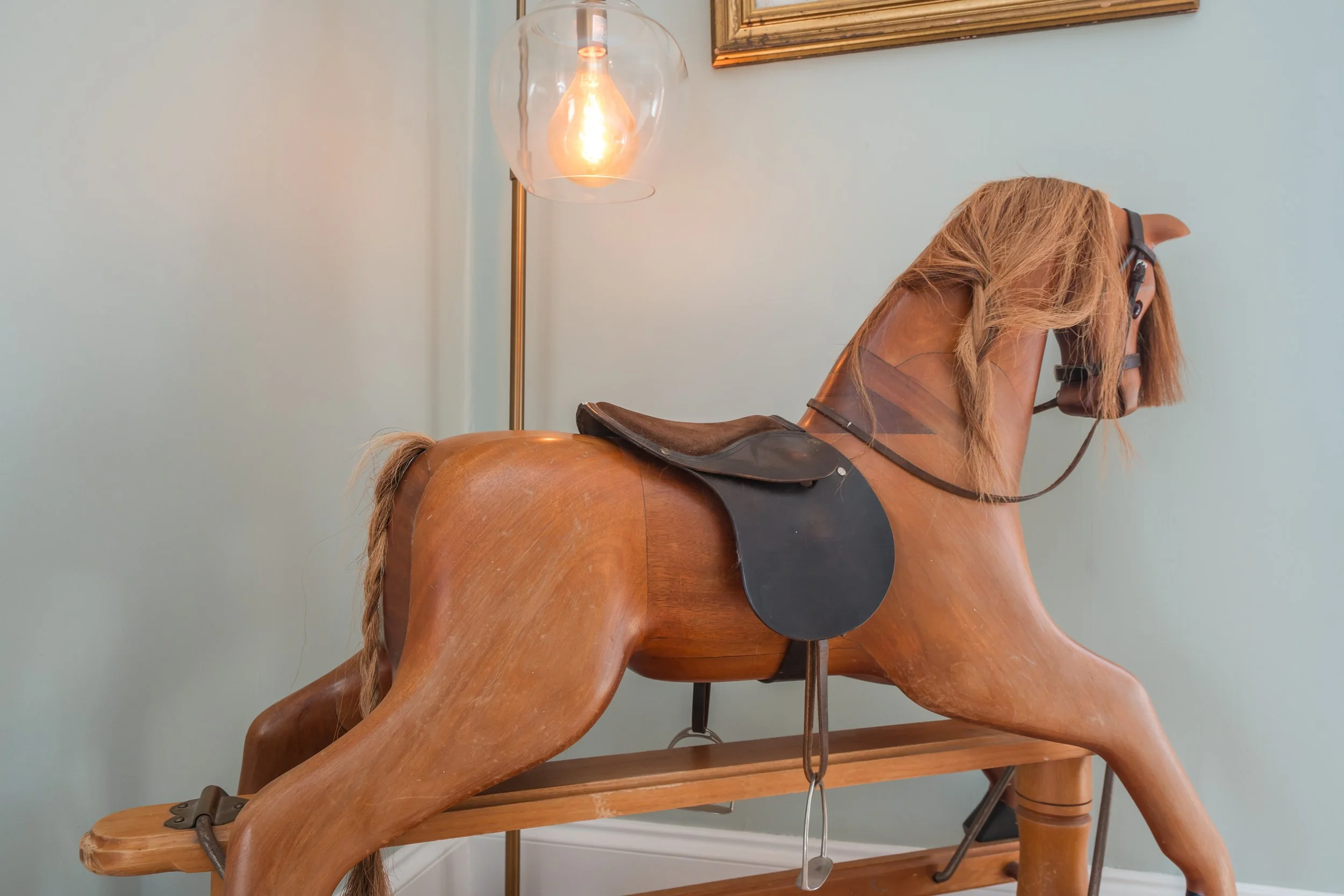 A wooden rocking horse with a saddle and bridle, positioned indoors near a light blue wall and a floor lamp with an exposed lightbulb.