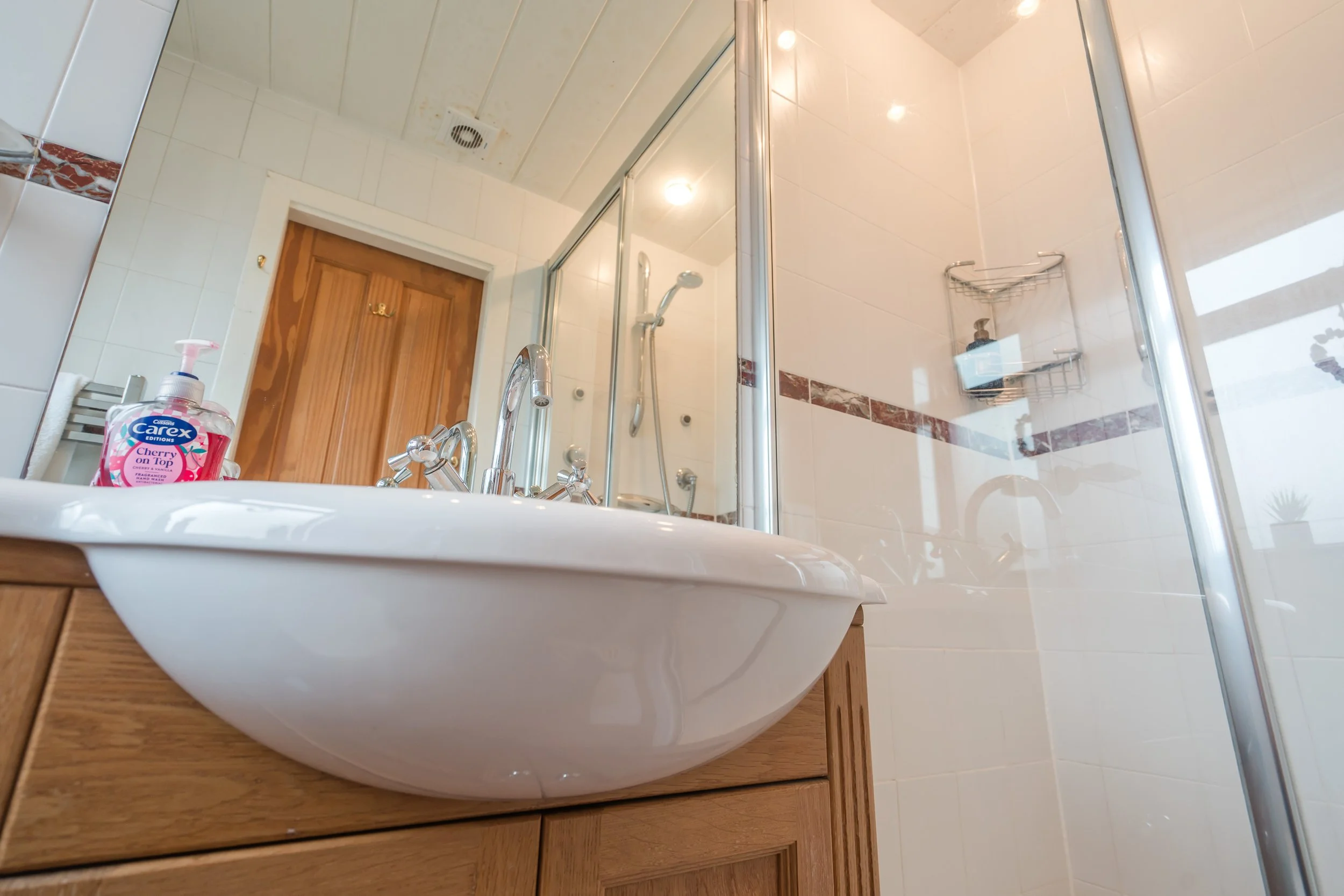 A bathroom with a sink, wooden cabinet, shower enclosure with glass door, white tiled walls with brown decorative stripe, and a wooden door in the background.