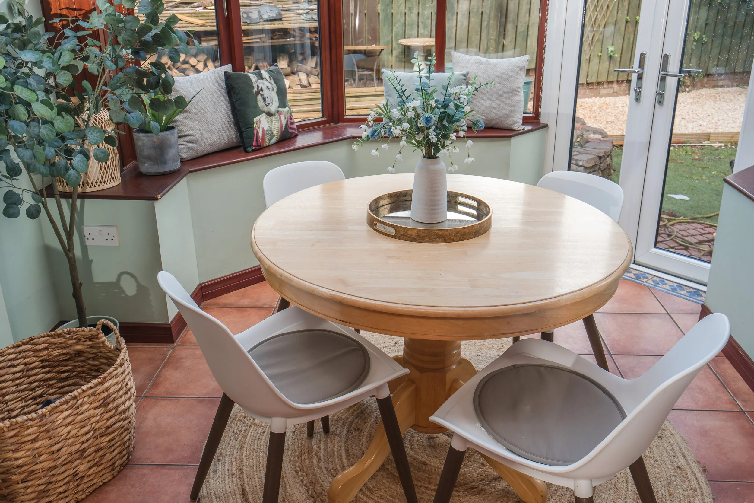 Dining area with a round wooden table, white chairs, a flower vase, and decorative pillows on window seat in sunlit conservatory