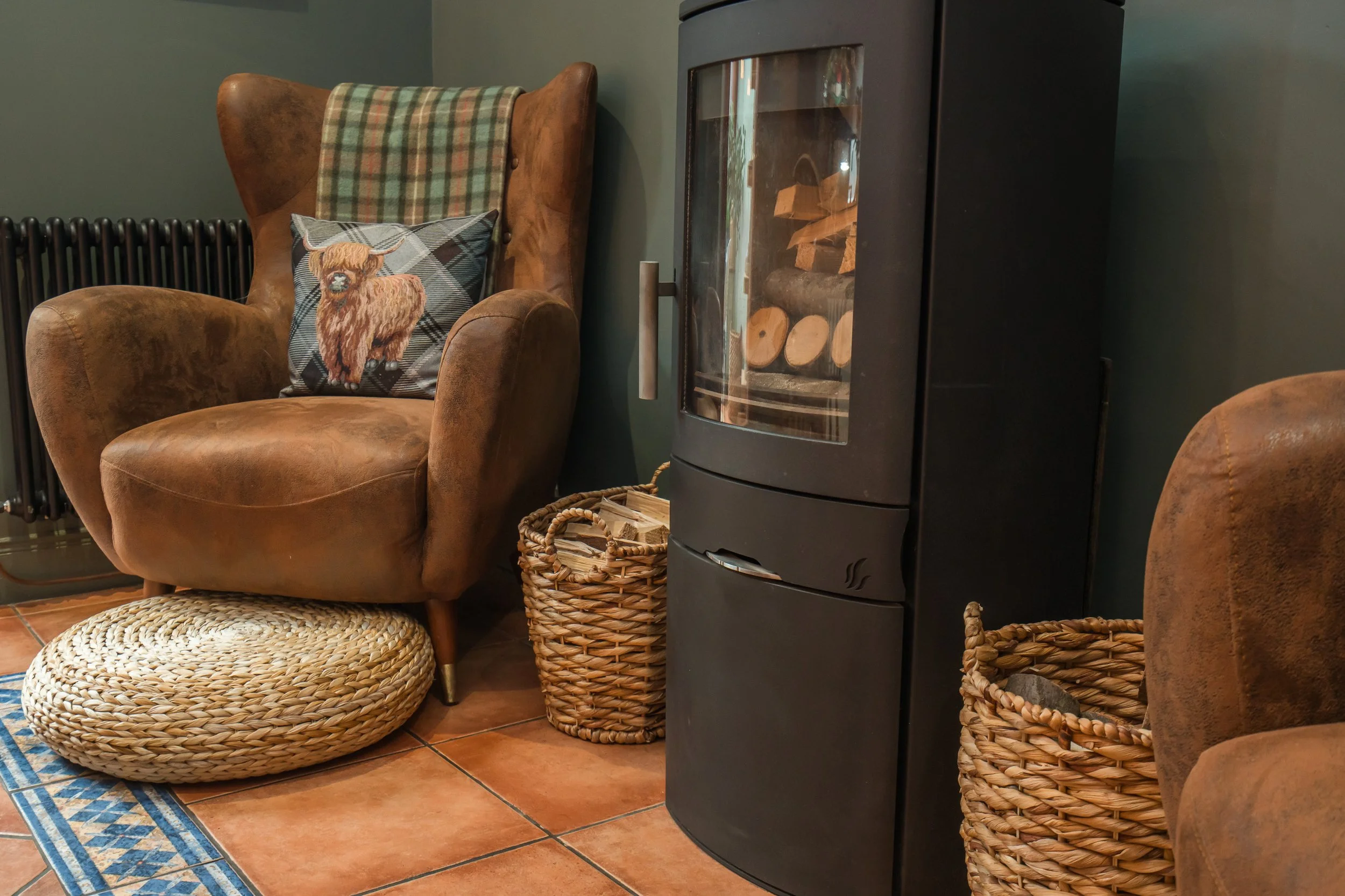 Cozy living room corner with an armchair holding a decorative pillow with a Highland cow design, a wicker basket with firewood, a black wood stove with glass door, and woven footstools on a patterned rug.